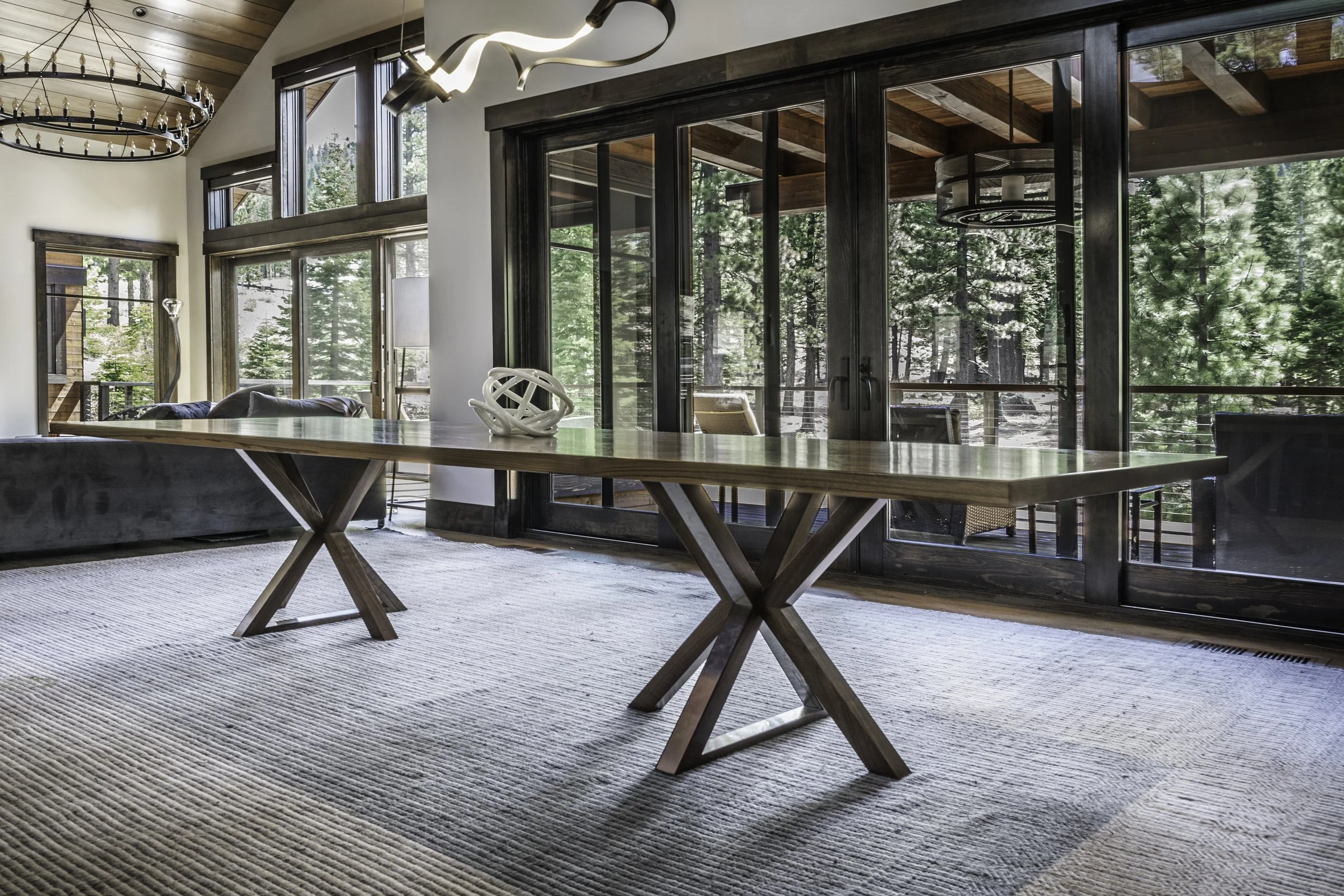 Living room with large windows and sliding glass doors showing a forest outside. Features a wooden table with a sculptural centerpiece, a sofa, and a textured area rug.