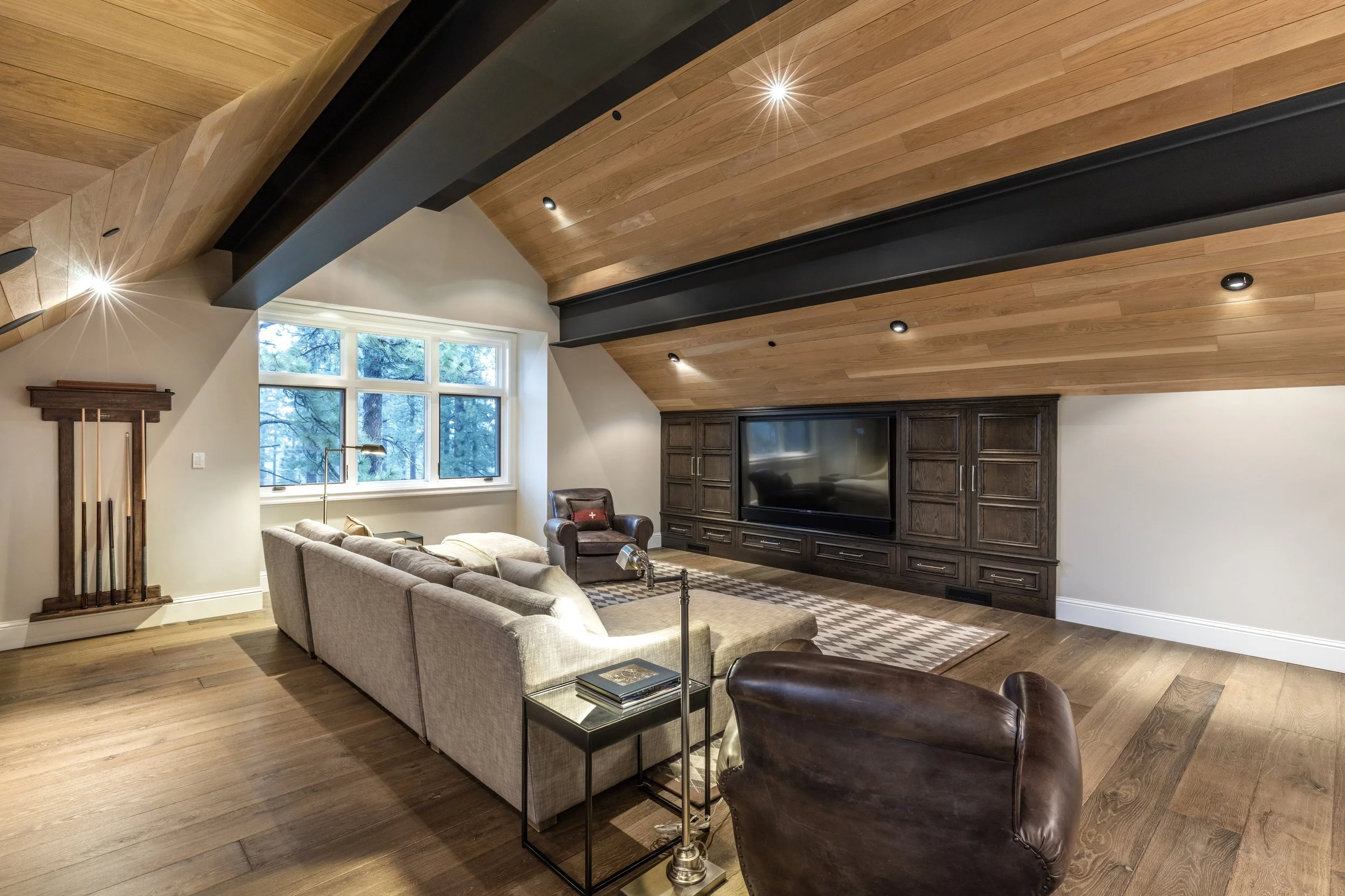 Living room with beige sectional sofa, leather armchair, wood-paneled sloped ceiling, large window, TV built into dark wood entertainment center, hardwood floors, and a patterned area rug.