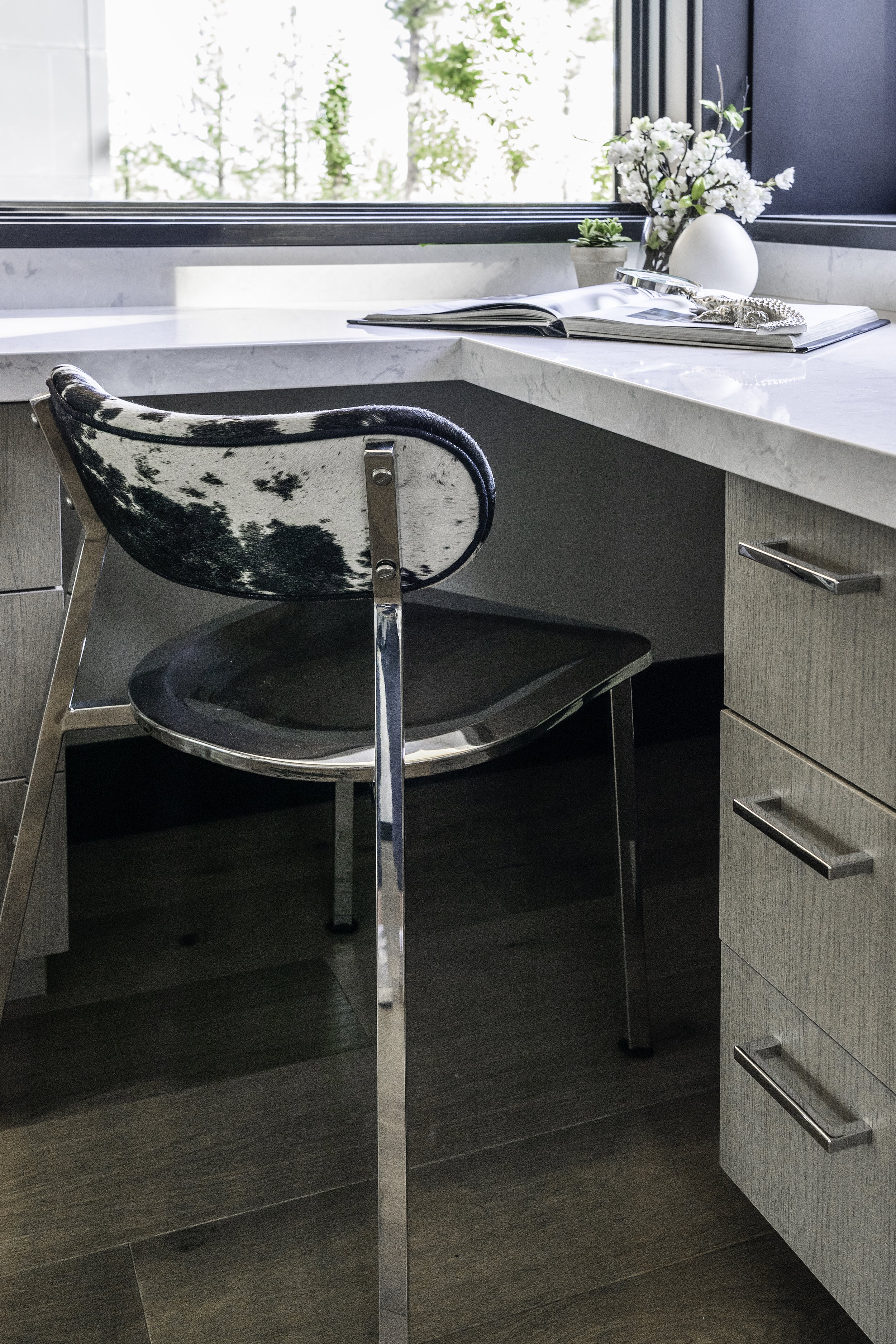 Modern office desk with a cowhide upholstered chair, a marble countertop, open magazine, outside view through window, decorative flowers, and potted plant.