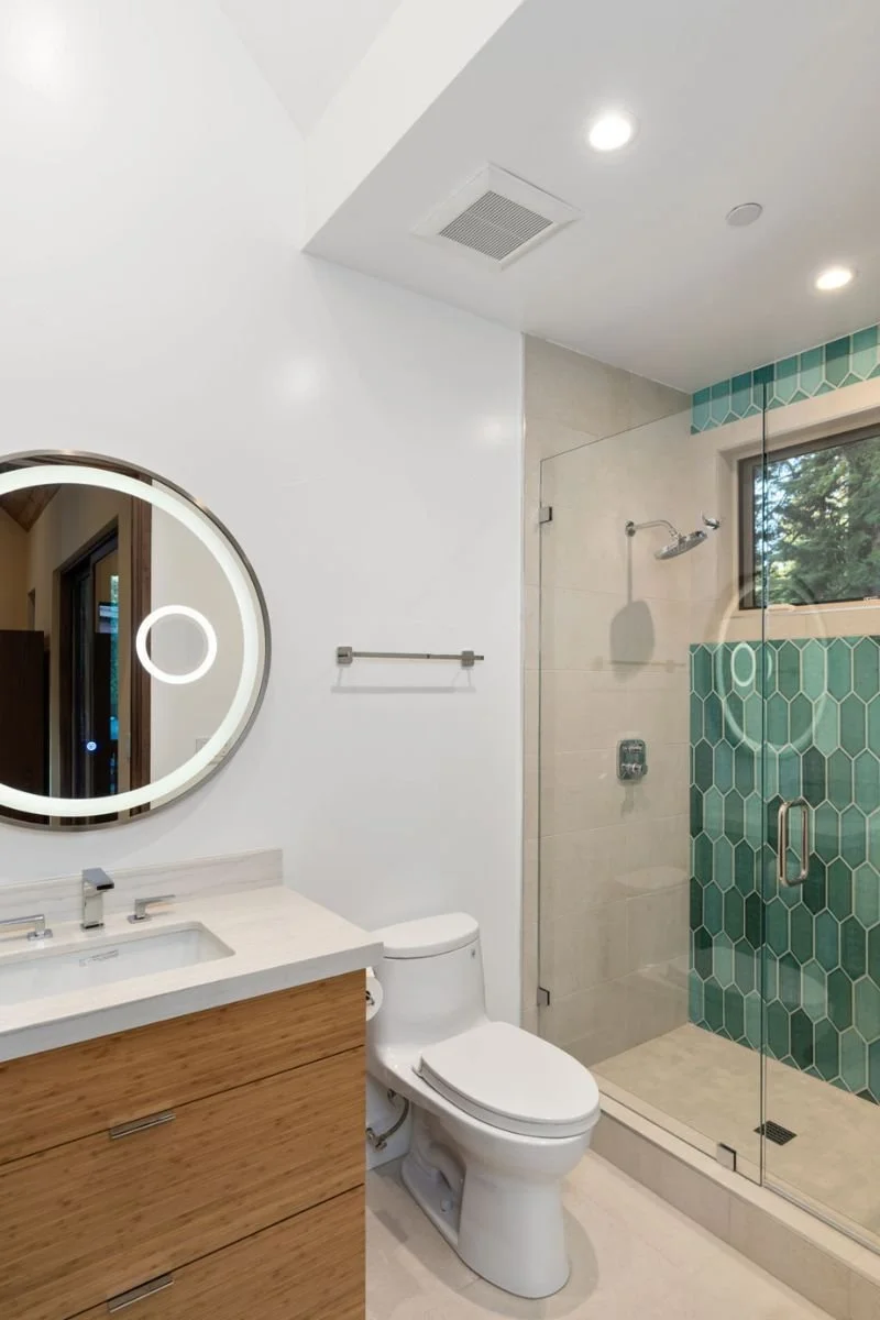 Modern bathroom with white walls, a round mirror with integrated LED lighting, a wooden vanity with a white countertop, a toilet, and a glass-enclosed shower with teal hexagonal tiles and a window showing trees outside.