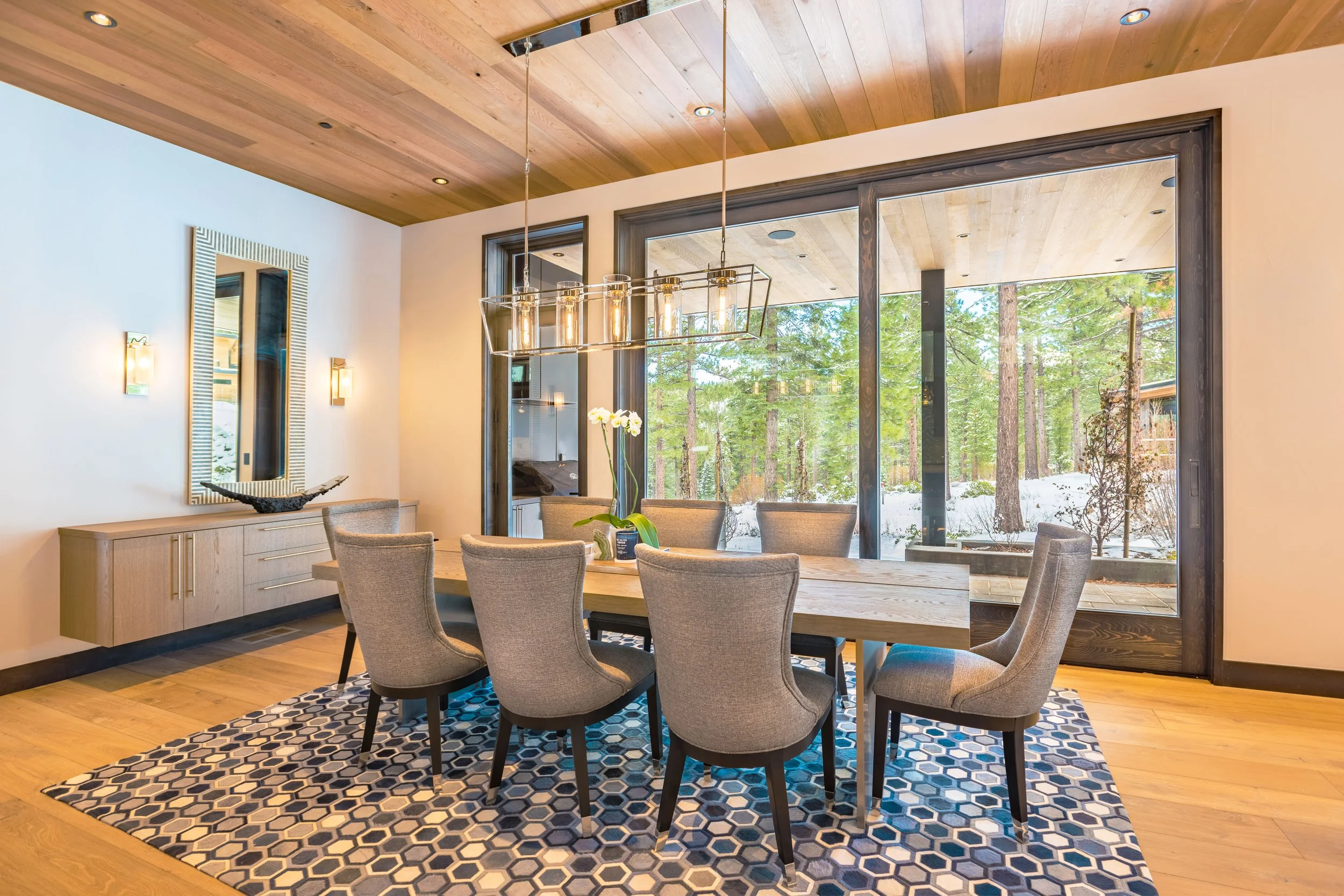 Modern dining room with a wooden table, eight upholstered chairs, a hexagonal patterned rug, large glass sliding doors showing a wintery forest outside, and a wooden ceiling with recessed lighting.