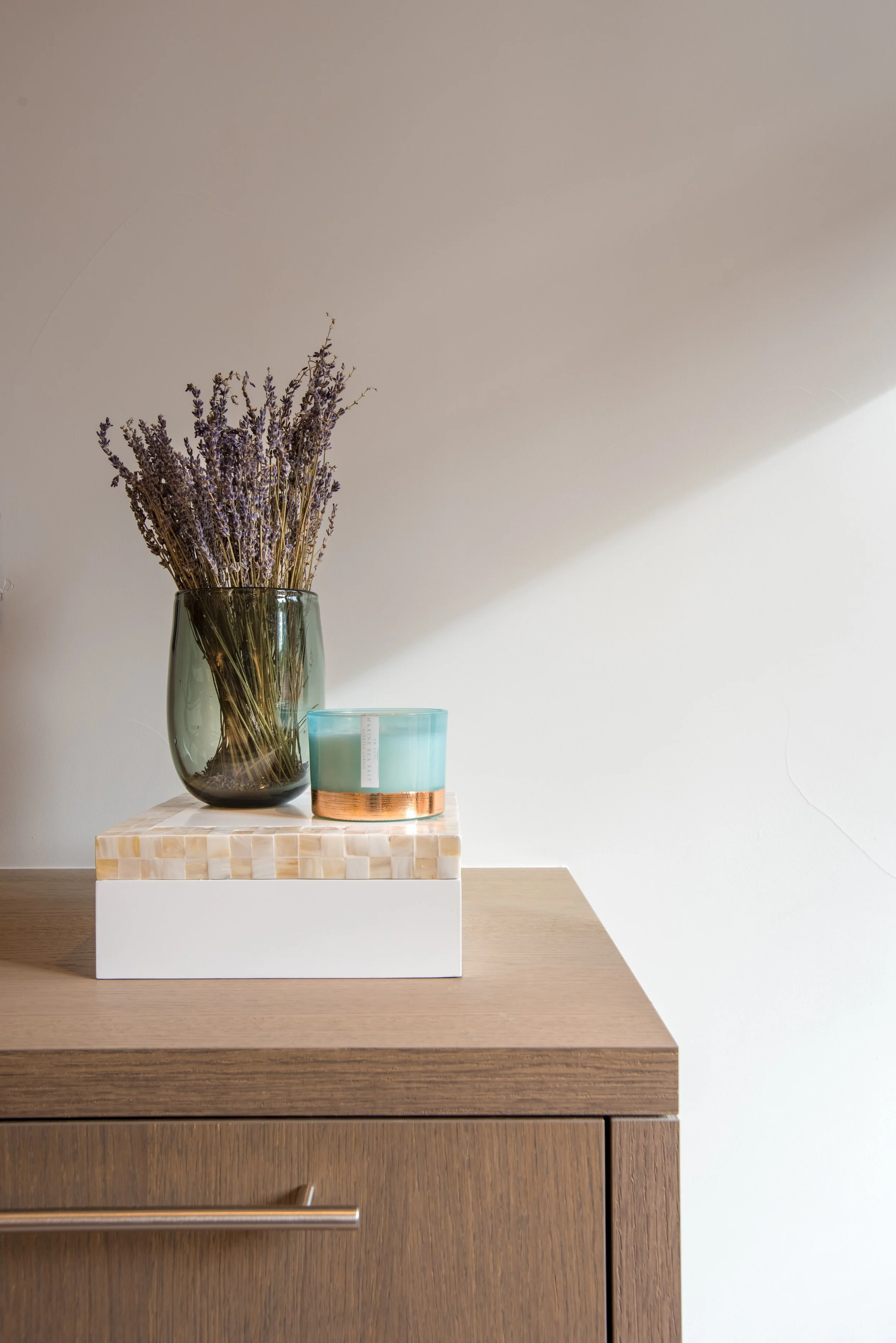 A wooden cabinet with a white top, featuring a green glass vase with dried lavender and a blue candle with a copper accent on a decorative white and beige tile platform.