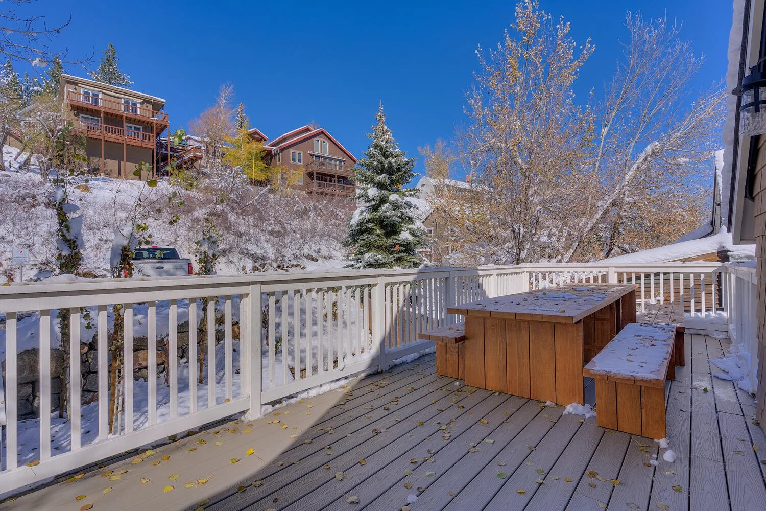 Snow-covered wooden deck with a picnic table and benches, surrounded by a white railing, overlooking a snowy landscape with trees and houses on a hillside under a clear blue sky.