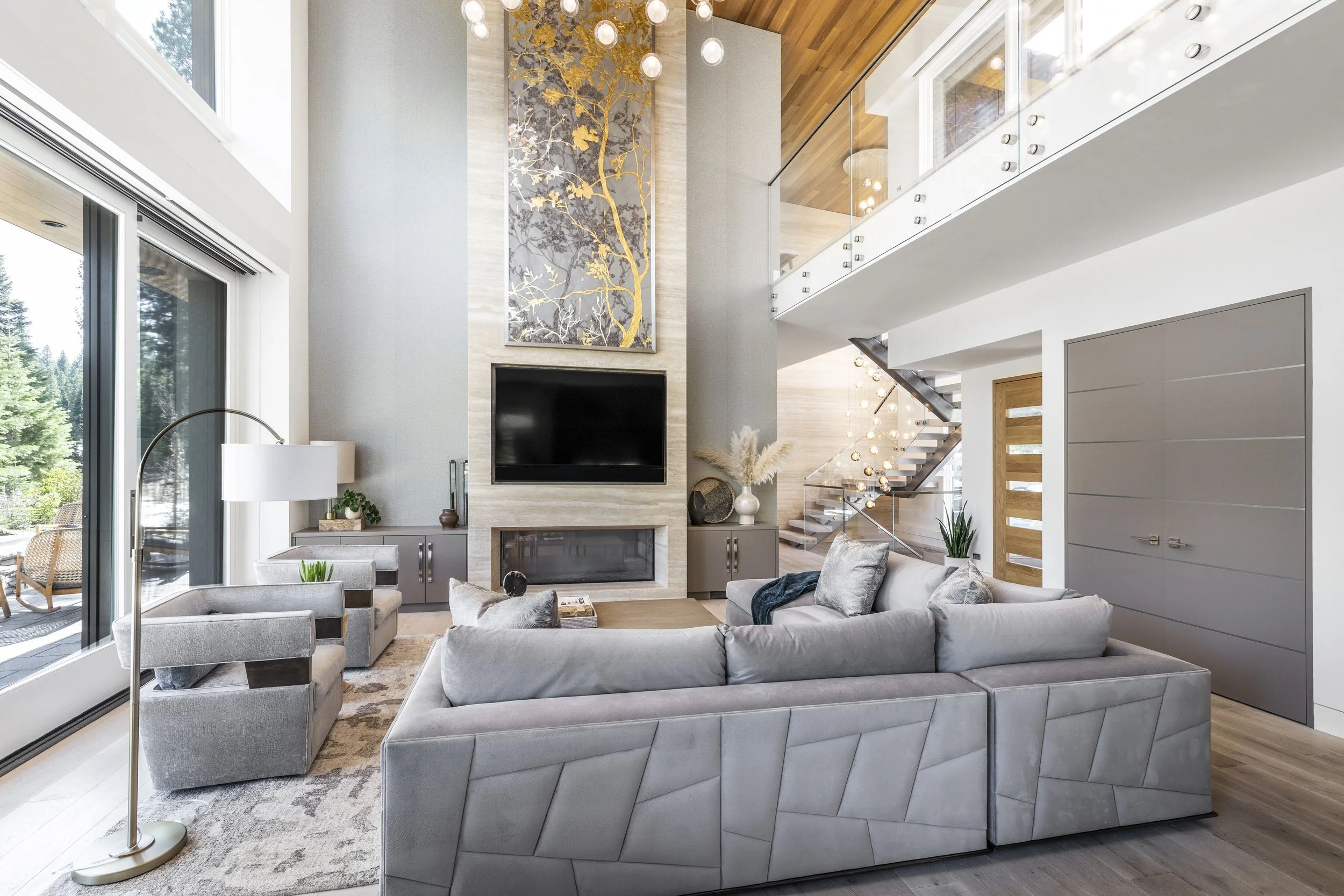 Modern living room with gray sectional sofa, two gray armchairs, a floor lamp, fireplace, wall-mounted TV, large windows, staircase, and decorative art and plants.