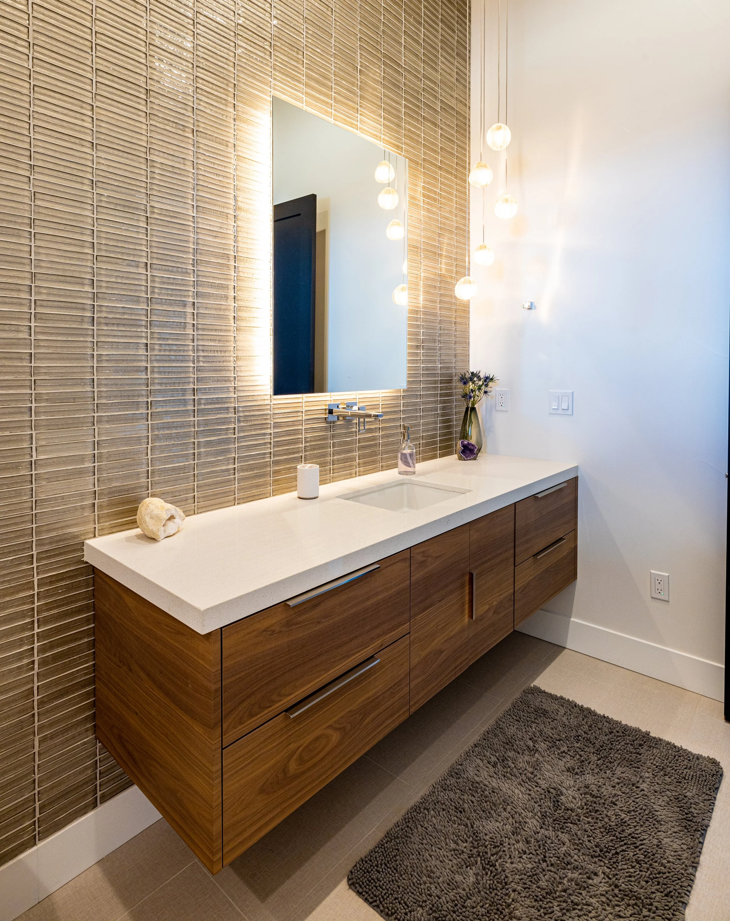 Modern bathroom vanity with a white countertop, wood cabinets, a large mirror, hanging globe lights, and decorative items, including a vase with flowers, a soap dispenser, and a rock.
