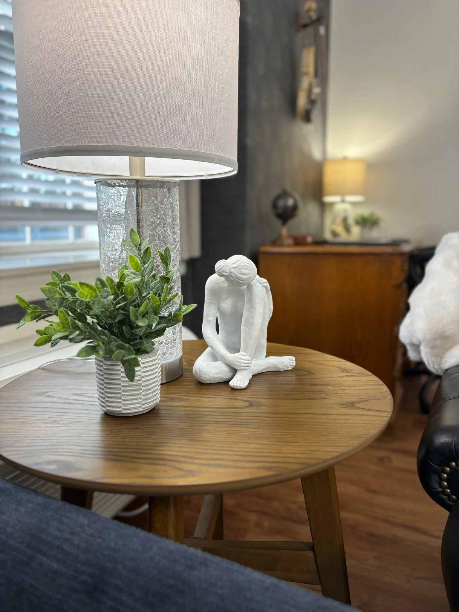 A round wooden side table with a white ceramic sculpture of a seated woman, a potted green plant, and a table lamp with a white shade. Make It Mine Season 6 Episode 2 show-Brookdale Senior Living.