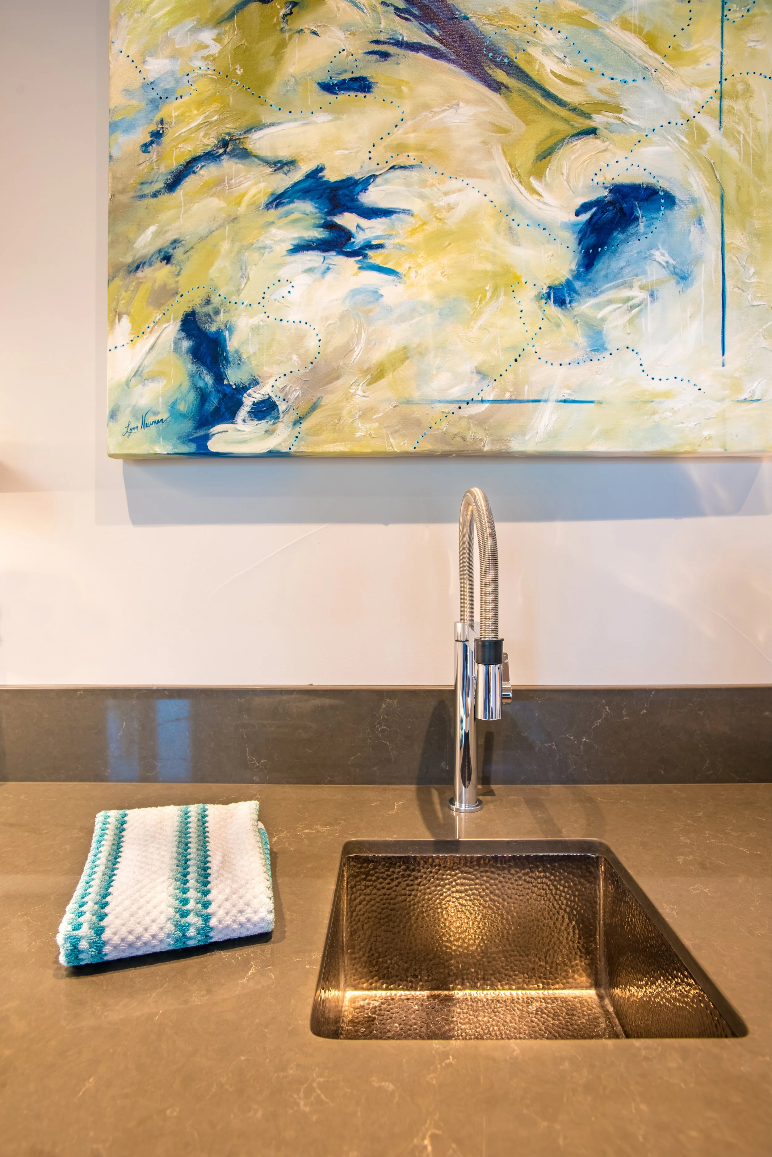 A kitchen or bar area with a dark stone countertop, a stainless steel sink, a modern chrome faucet, a folded white dish towel with blue stripes, a white wall, a colorful abstract painting with blue, yellow, and white brushstrokes hanging above the si