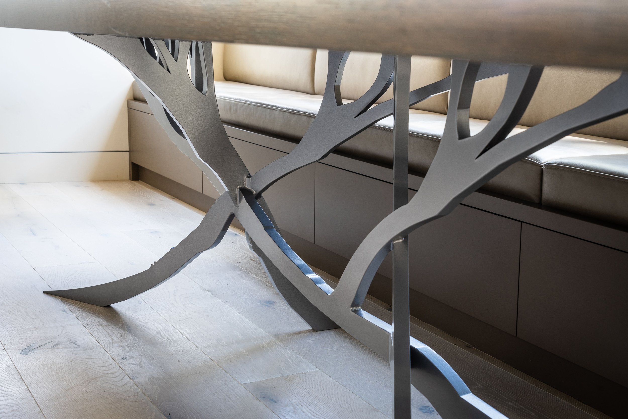 Close-up of a modern metal chair leg with a tree branch shape, positioned underneath a wooden table. In the background, there is a built-in bench with beige cushions and light wood flooring.