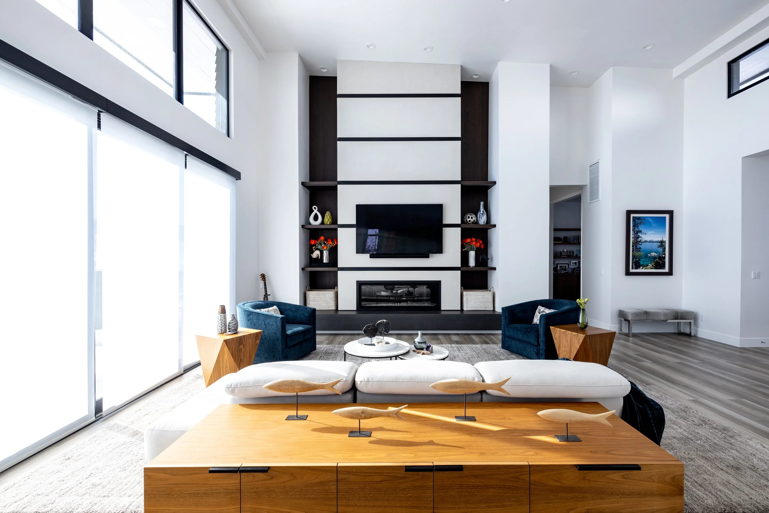 Modern living room with a fireplace, mounted TV, and built-in shelves, featuring two blue armchairs, a white couch with fish sculptures on a wooden table, and large sliding glass doors.