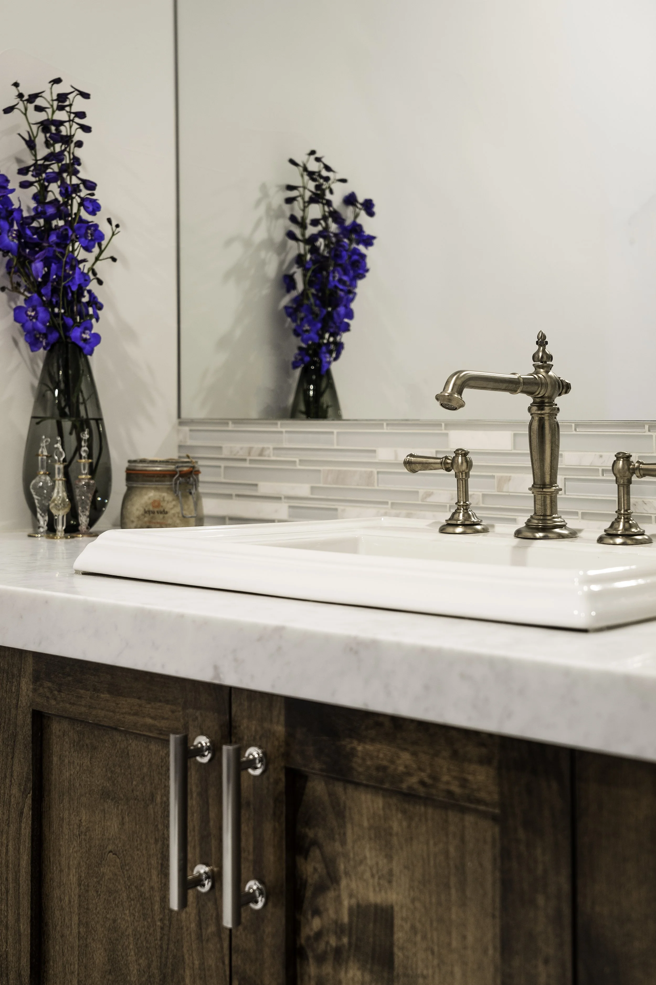 Bathroom sink with metallic faucet, white countertop, wooden cabinetry, and blue flower vases reflected in mirror.