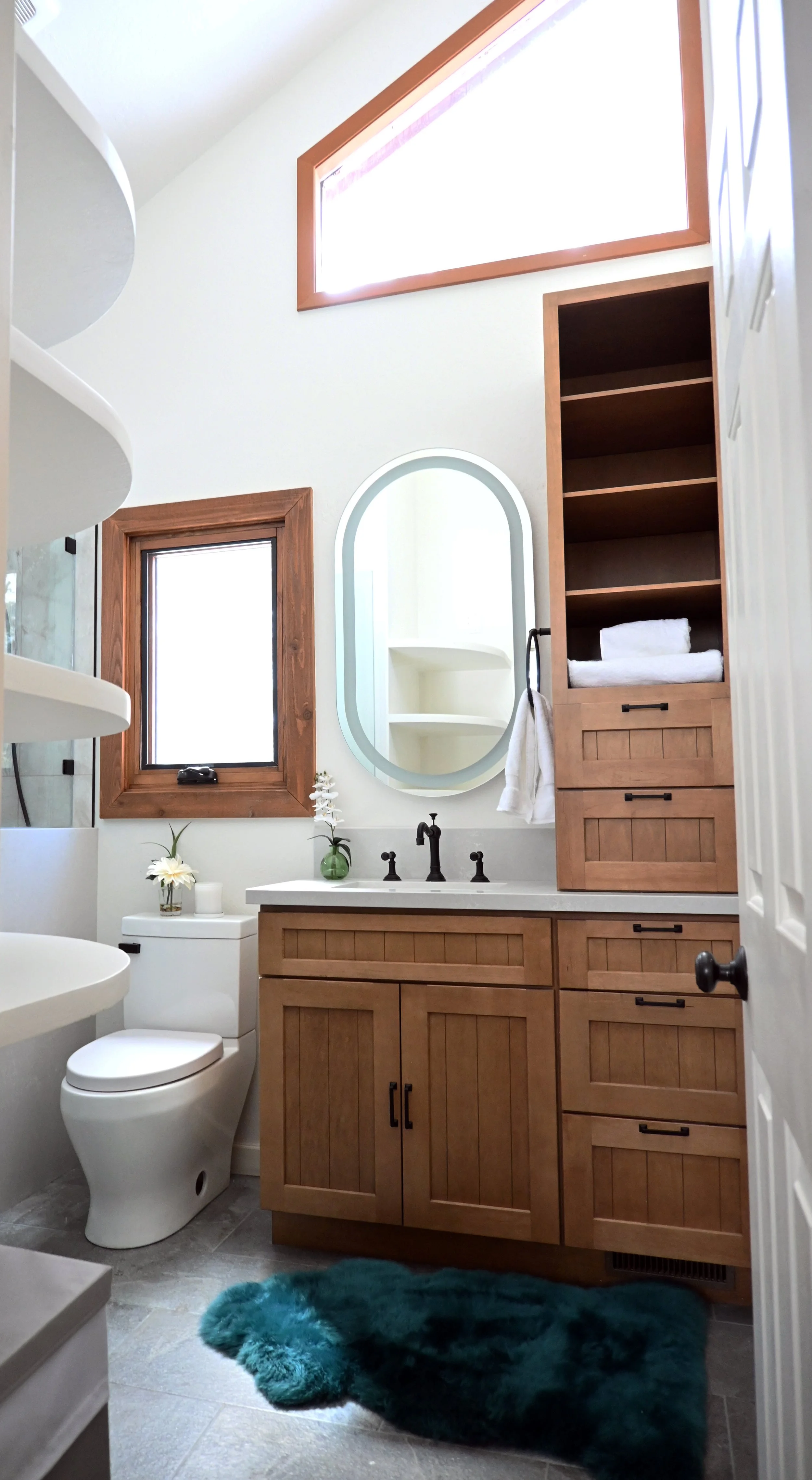 Bathroom with wooden cabinetry, an oval mirror, small window, teal rug, and decorated with a vase of white flowers.