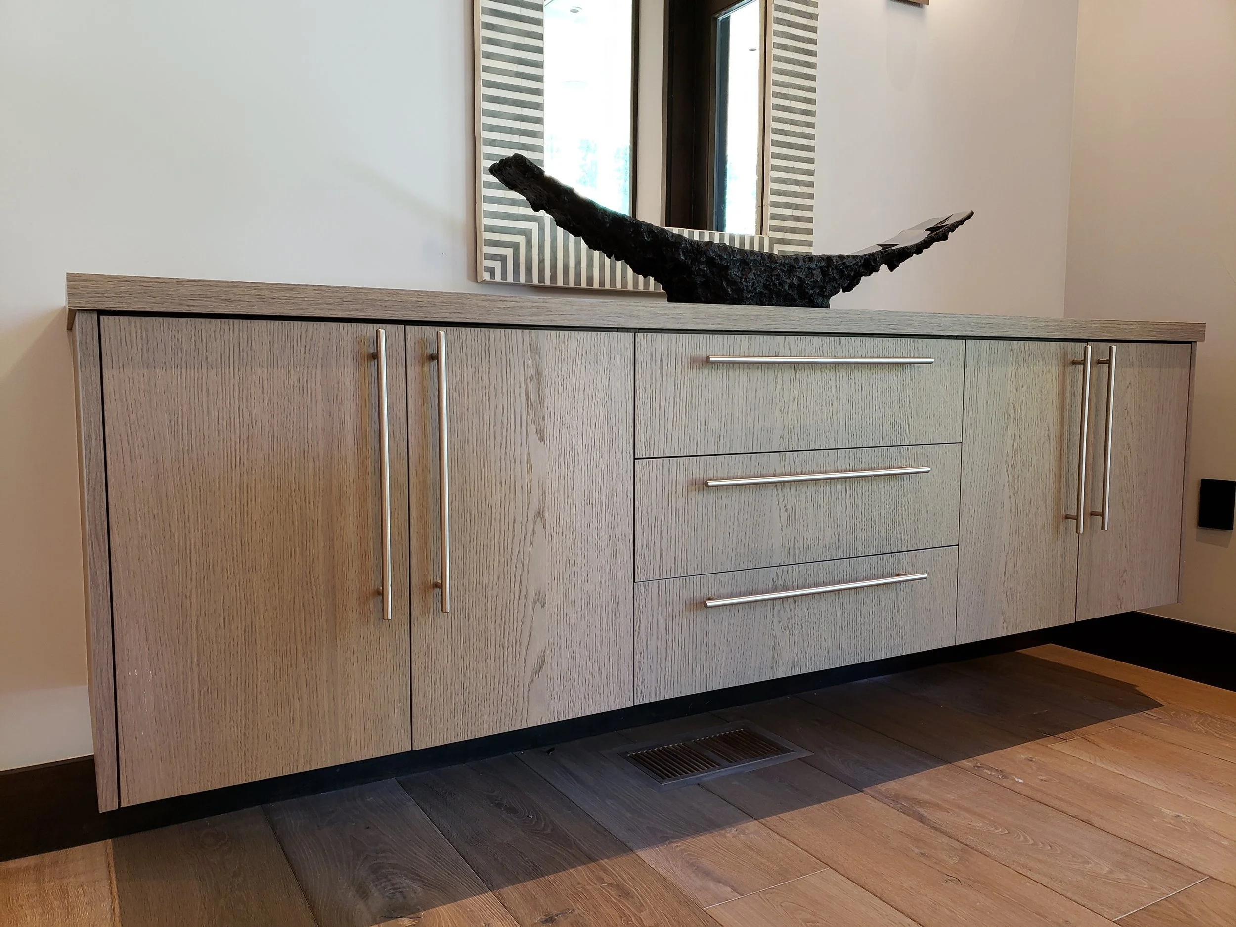 A wooden sideboard with three drawers and two cabinets, metal handles, placed against a wall. There is a large mirror and a black decorative sculpture on top. The floor is wood, and a vent is visible underneath the sideboard.