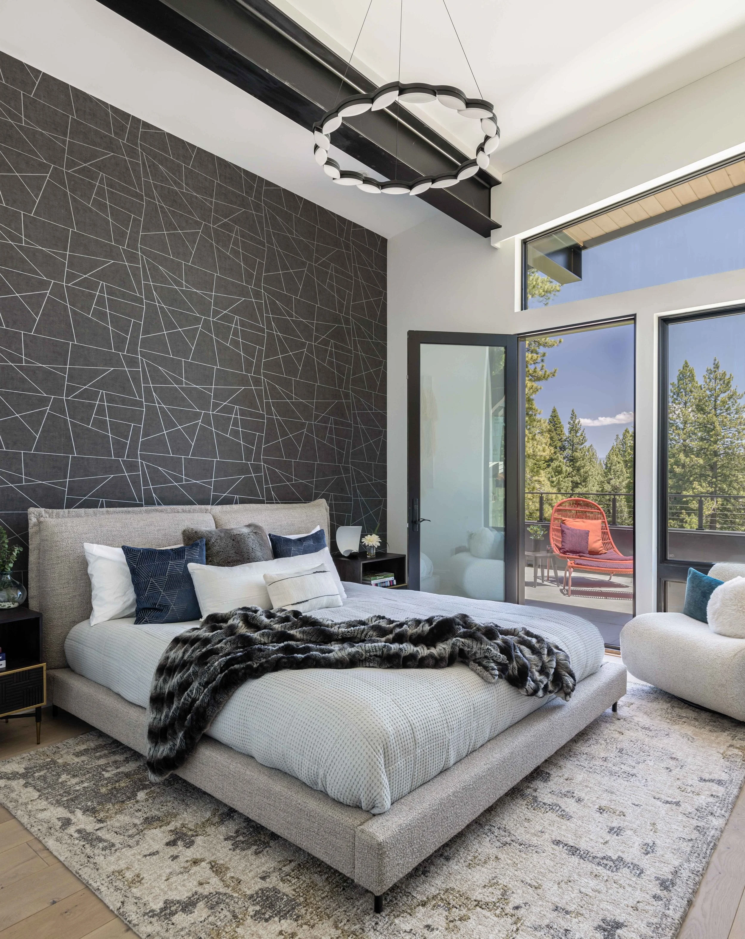 Modern bedroom with a large bed, dark geometric accent wall, black nightstands, and large windows overlooking a balcony with outdoor furniture and trees.