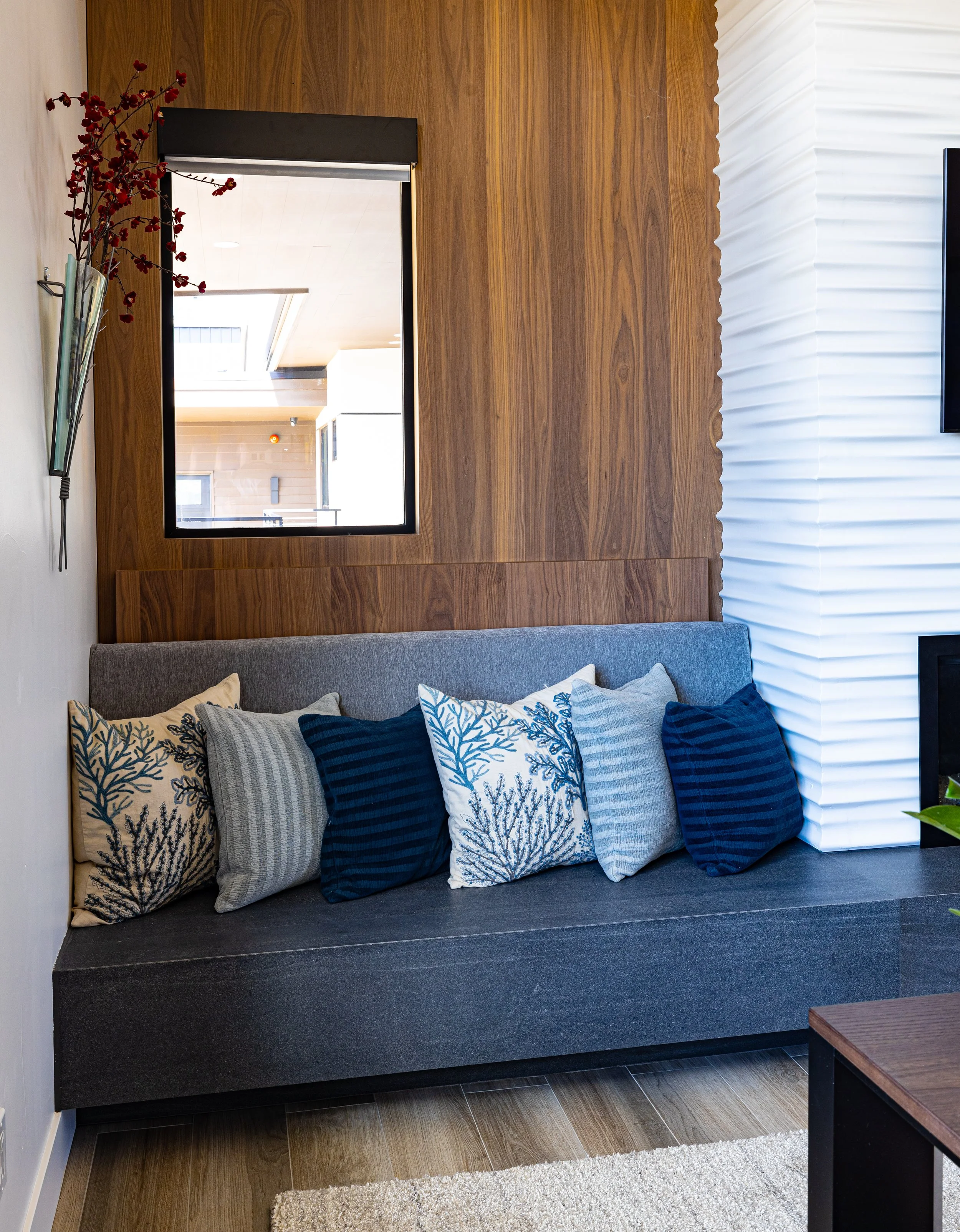 Living room with a gray sofa adorned with six decorative pillows in shades of blue, gray, and beige, against a paneled wood wall. A large mirror is on the wall above the sofa, reflecting part of the ceiling and windows. To the left, there is a wall-m