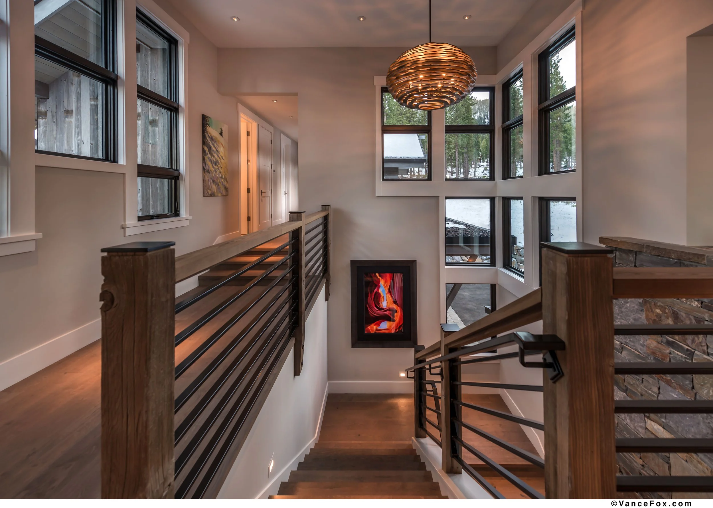 Interior view of a staircase landing in a modern house with large windows, wooden handrail, framed artwork, a fireplace, and a view of snow outside.