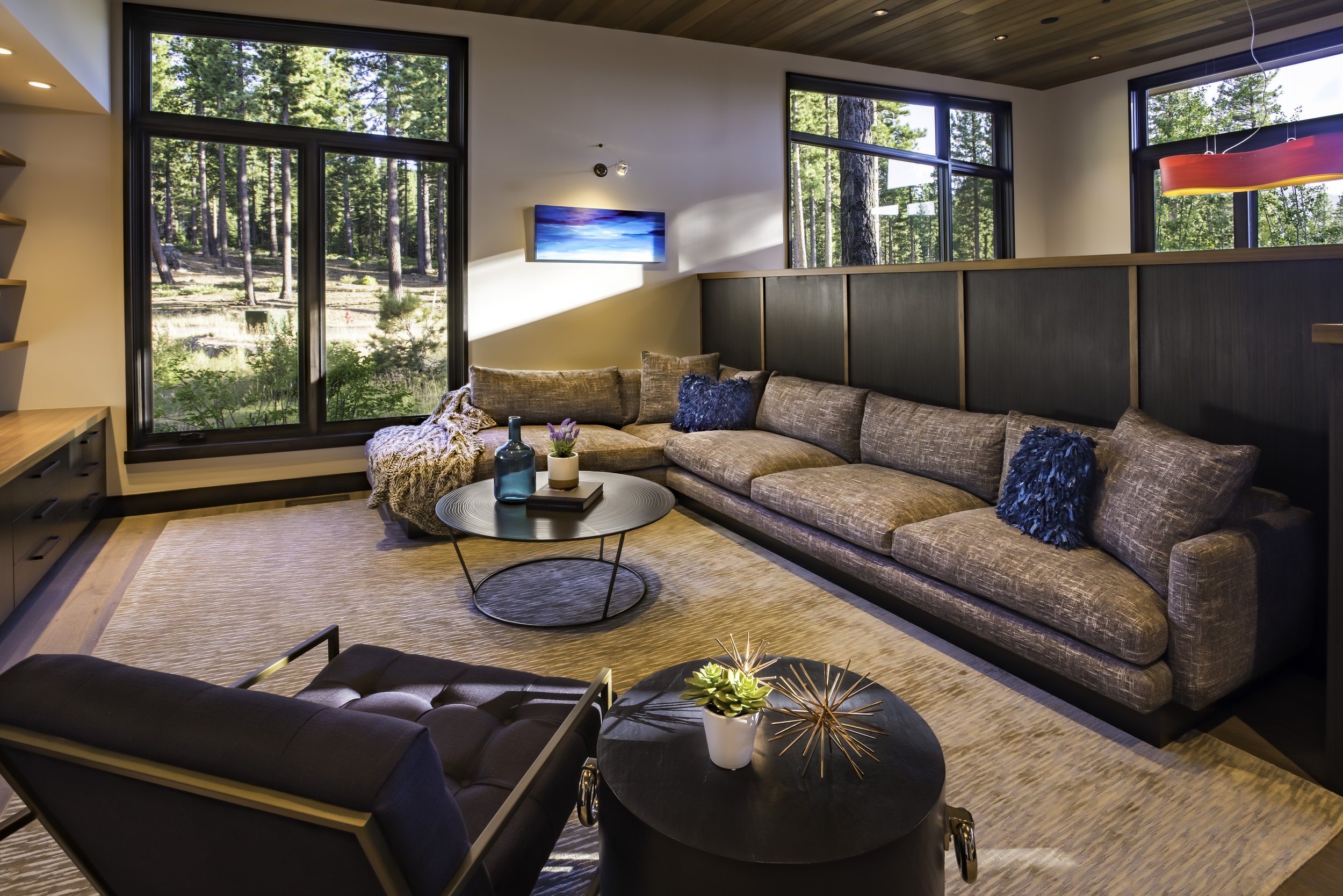 Living room with large windows showcasing a forest view, brown sectional sofa with blue and beige pillows, round coffee table with vases and plants, and modern decor.