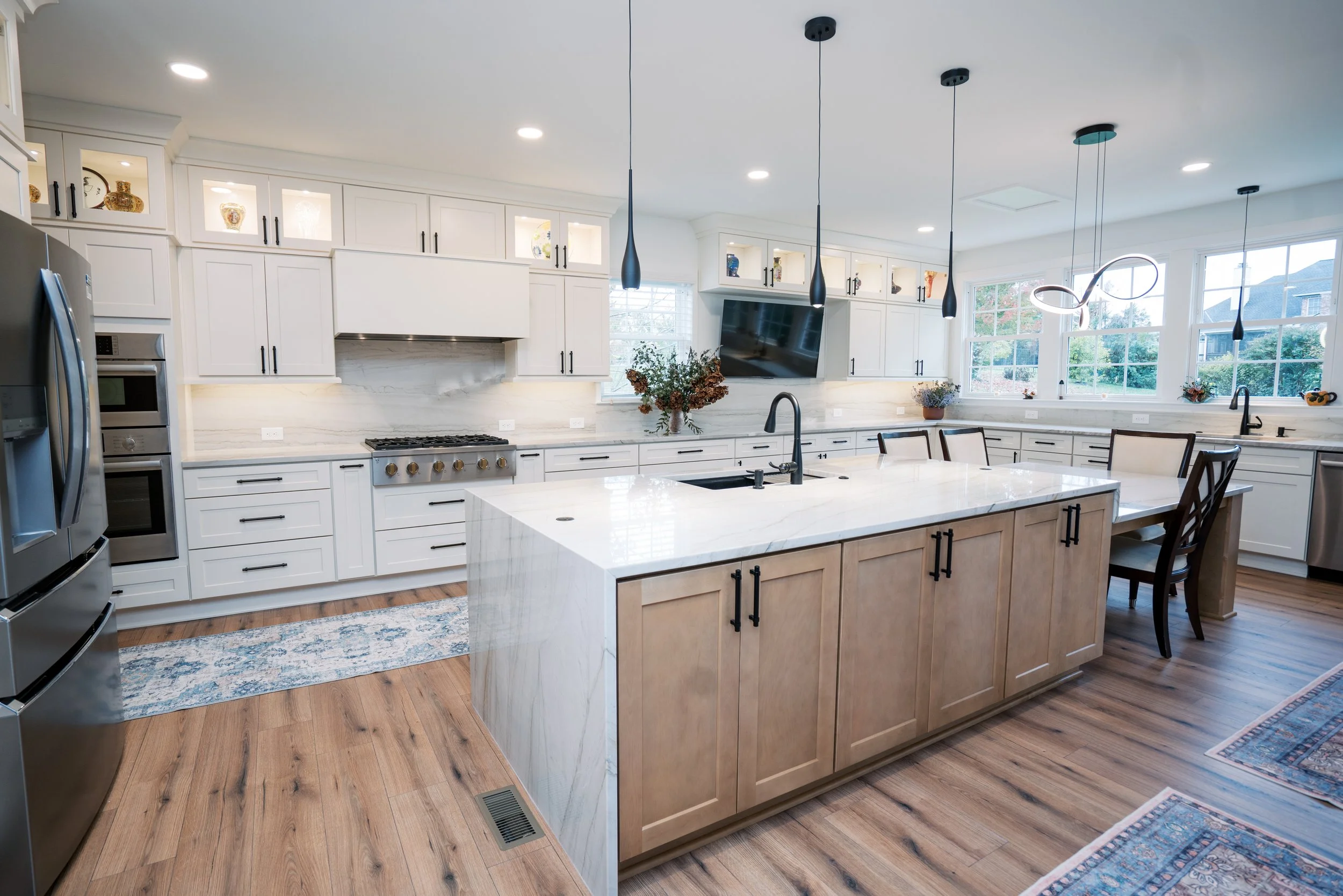 Modern kitchen with white cabinetry, a large central island with a marble countertop, stainless steel appliances, a dining table with chairs, and large windows letting in natural light.