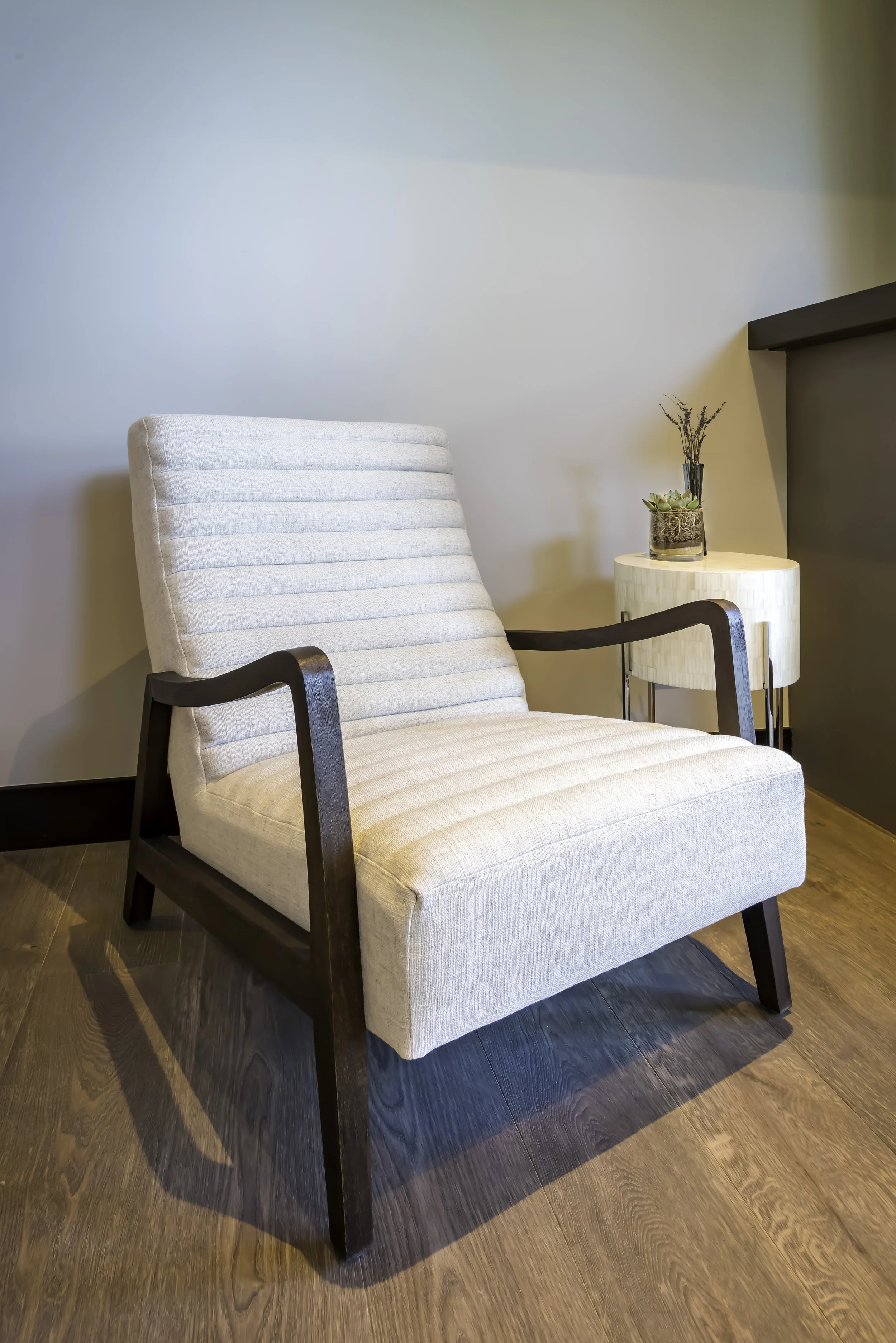 An beige upholstered armchair with dark wooden armrests and legs, placed next to a cream-colored side table with a potted succulent plant, on a wooden floor against a neutral wall.
