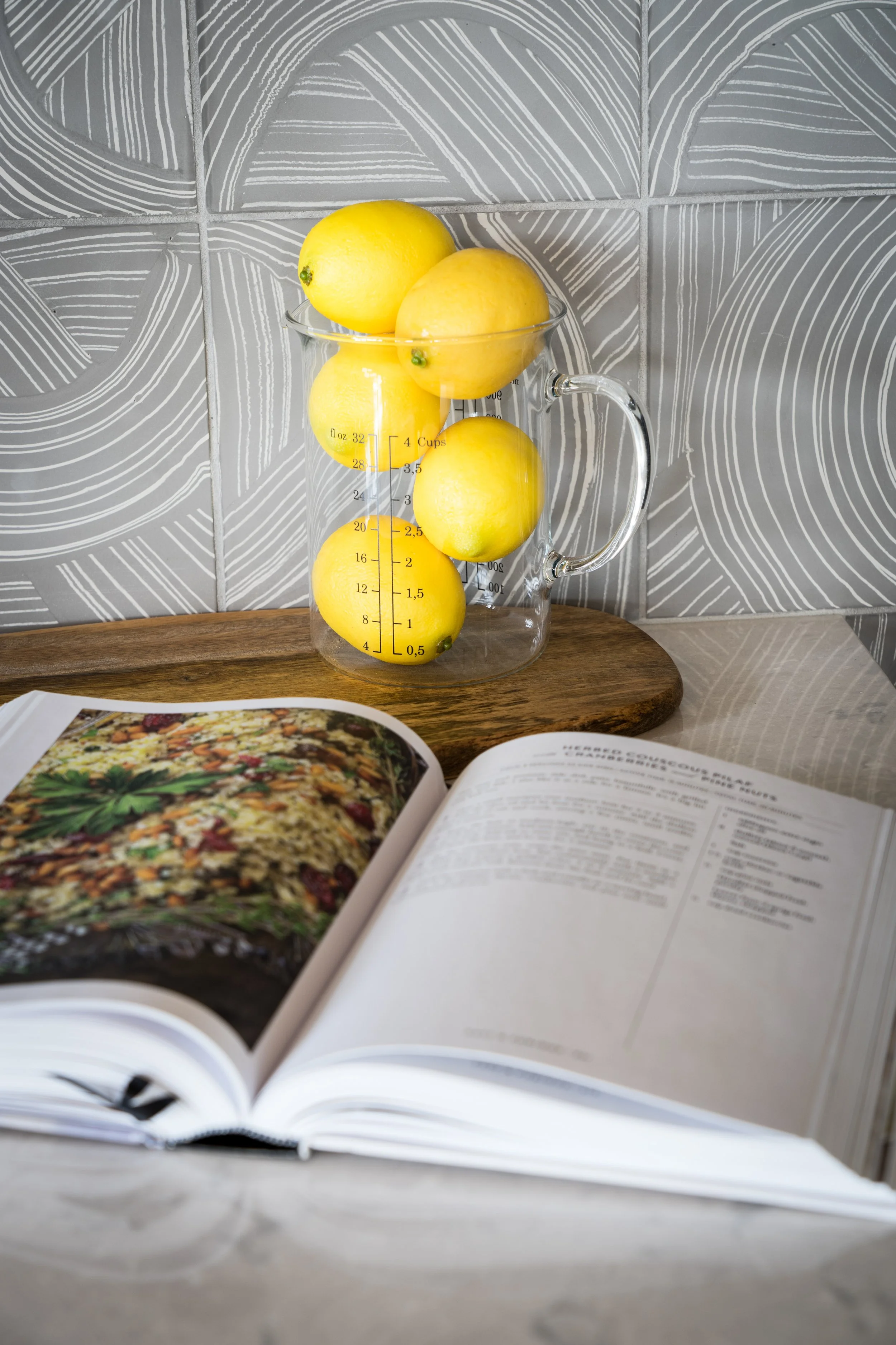 A glass measuring cup with lemons inside, placed on a wooden surface with an open cookbook in the foreground, and decorated gray tiles in the background.