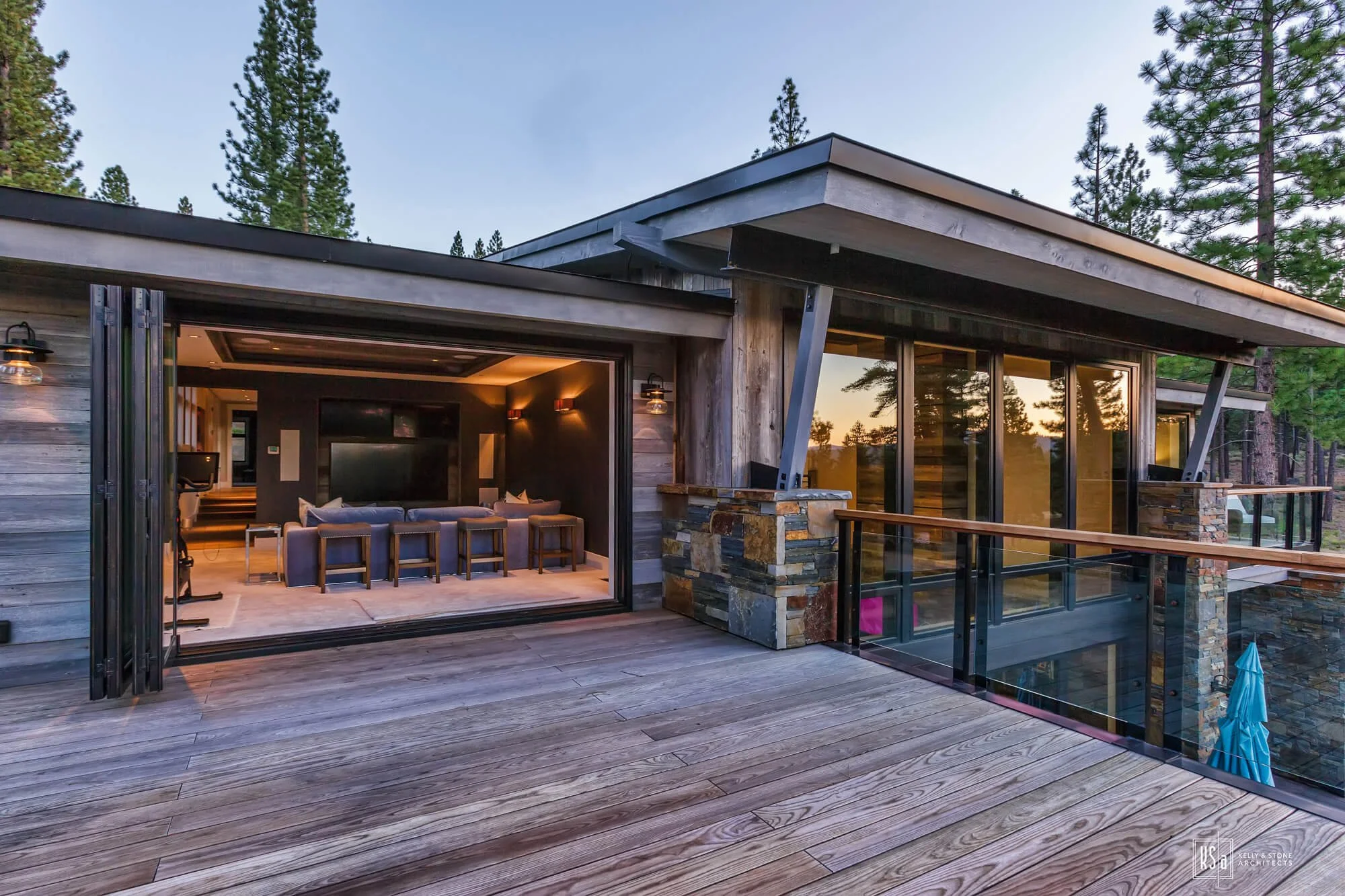Outdoor balcony of a modern house with glass railing, wooden flooring, and trees in the background at sunset