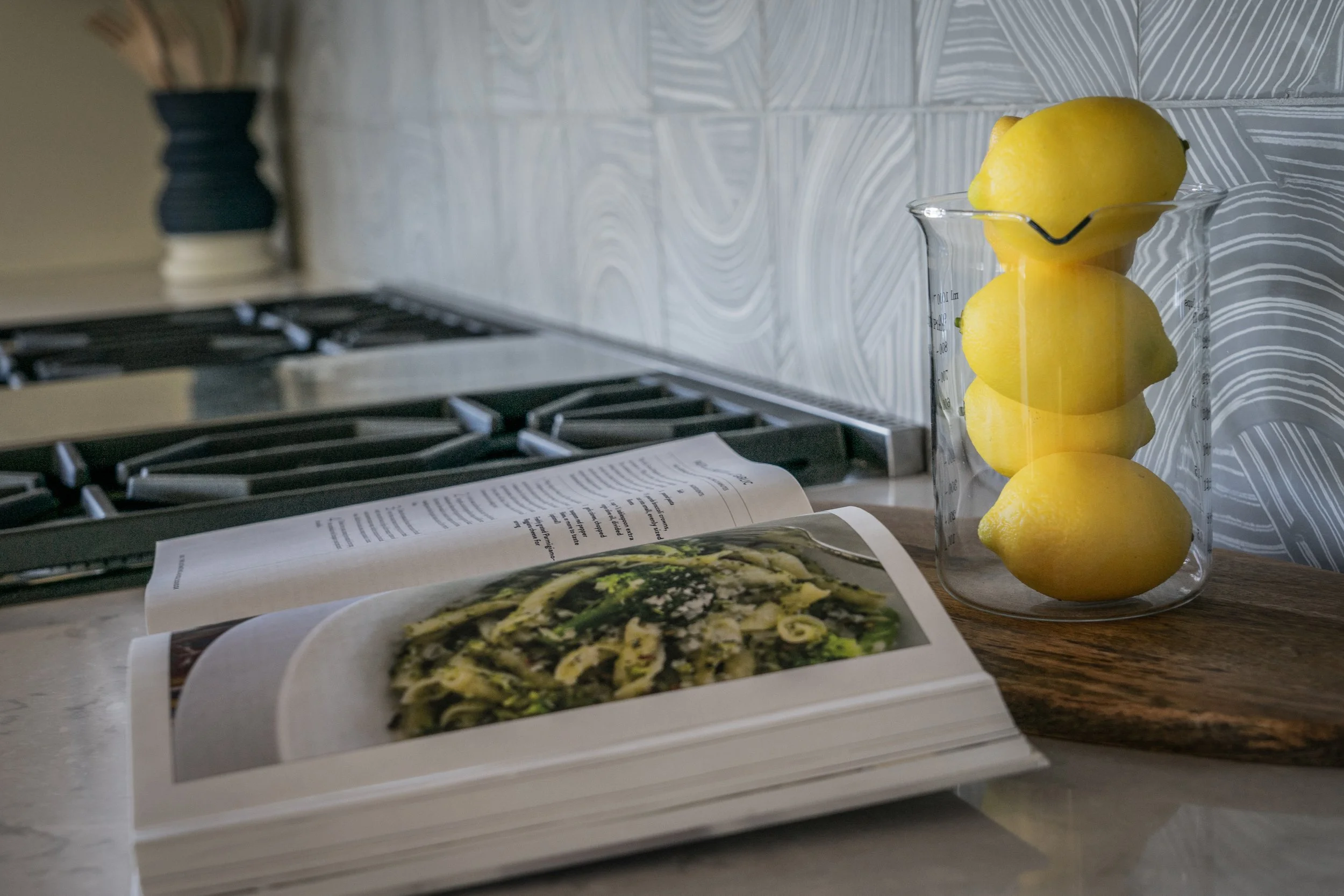 A glass jar with four lemons and an open cookbook displaying a photo of a green salad, all on a kitchen countertop with a stove in the background.