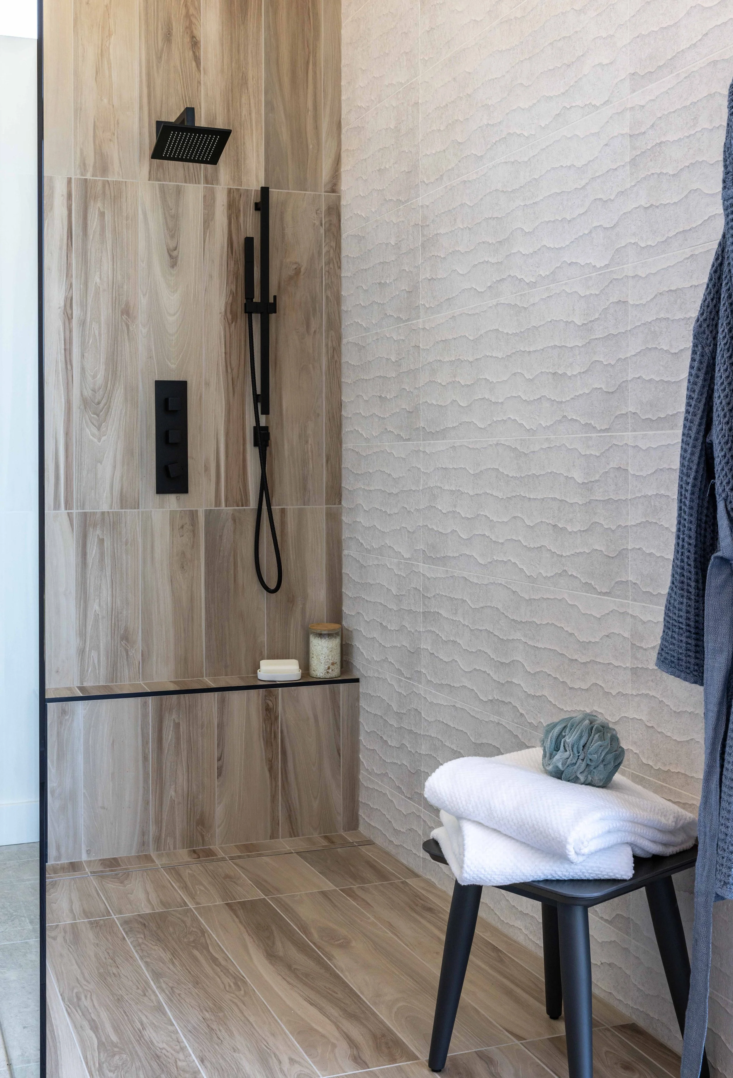 Modern shower with black fixtures, wood-look tiles, and textured wall, with a stool holding a towel and bath sponge.