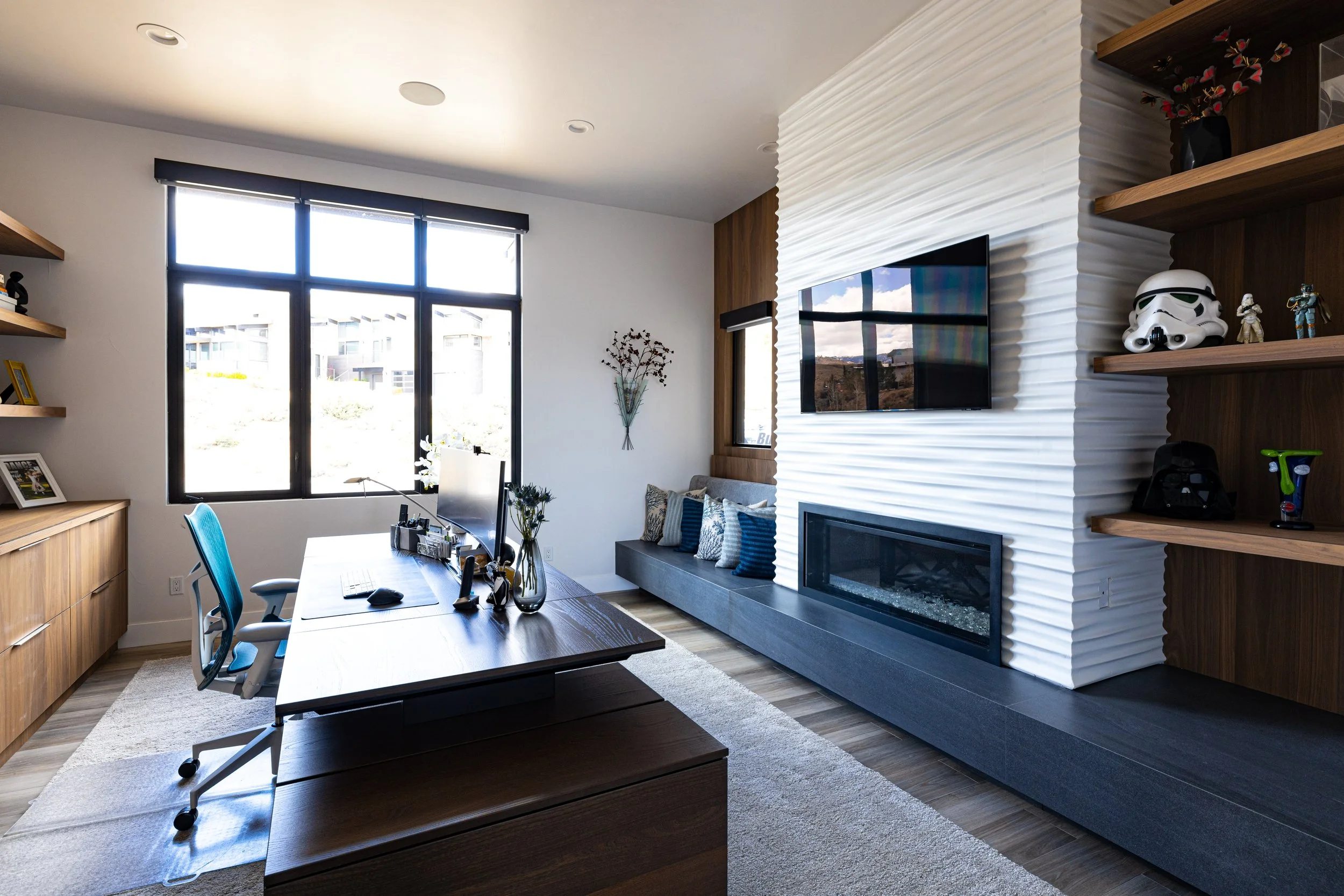 Modern home office with large window, wooden desk, blue and patterned pillows, white textured wall with fireplace and flat-screen TV, wooden shelving with Star Wars and other collectibles.