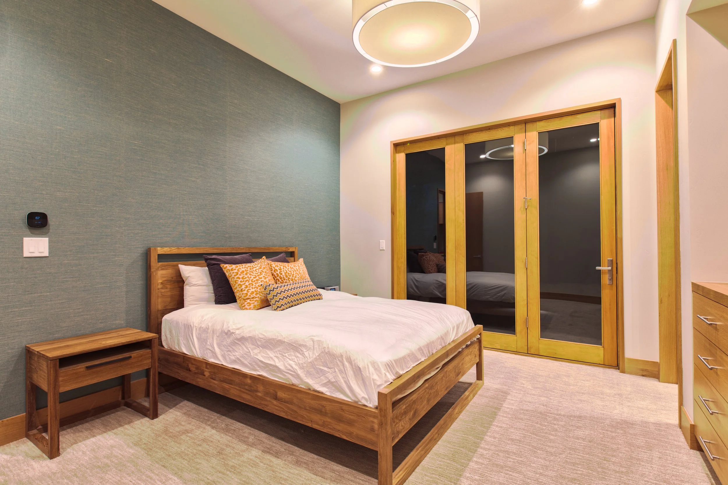 A modern bedroom with a wooden bed, colorful pillows, a nightstand, a textured green accent wall, and double glass doors leading outside, illuminated by a circular ceiling light.