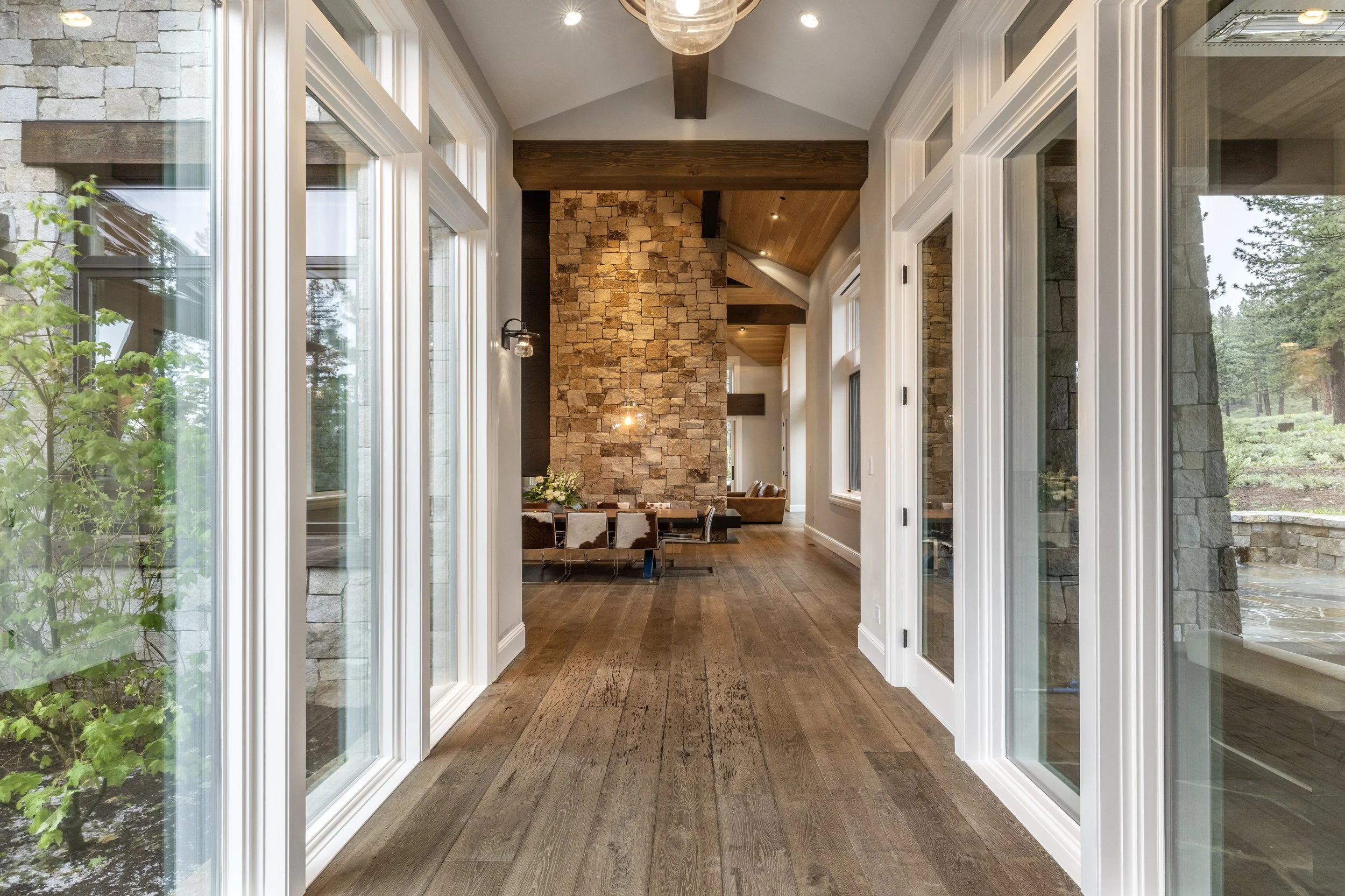 Bright hallway of a modern house with large windows, wooden floors, a stone fireplace wall, and glimpses of dining and living areas.