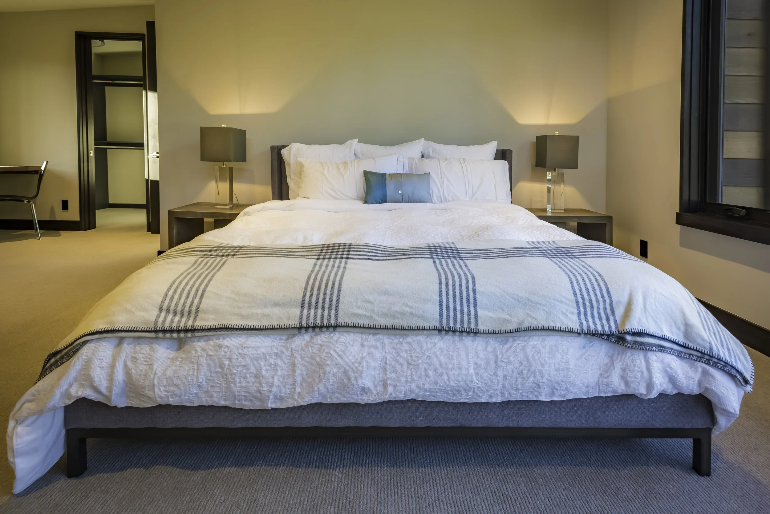Hotel room with a neatly made bed, white bedding, a small blue pillow, two nightstands with gray lamps, a window on the right, and a door leading to a closet or bathroom on the left.