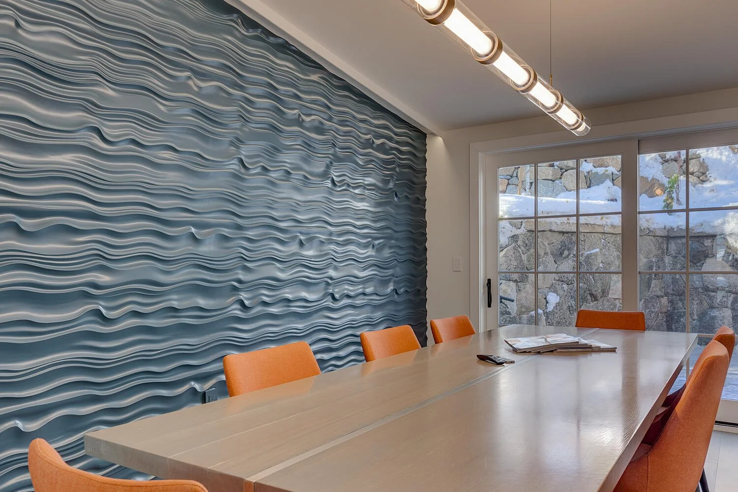 Interior of a modern dining room with a long table, orange chairs, a textured blue wall, and large glass doors showing a snowy stone outdoor scene.