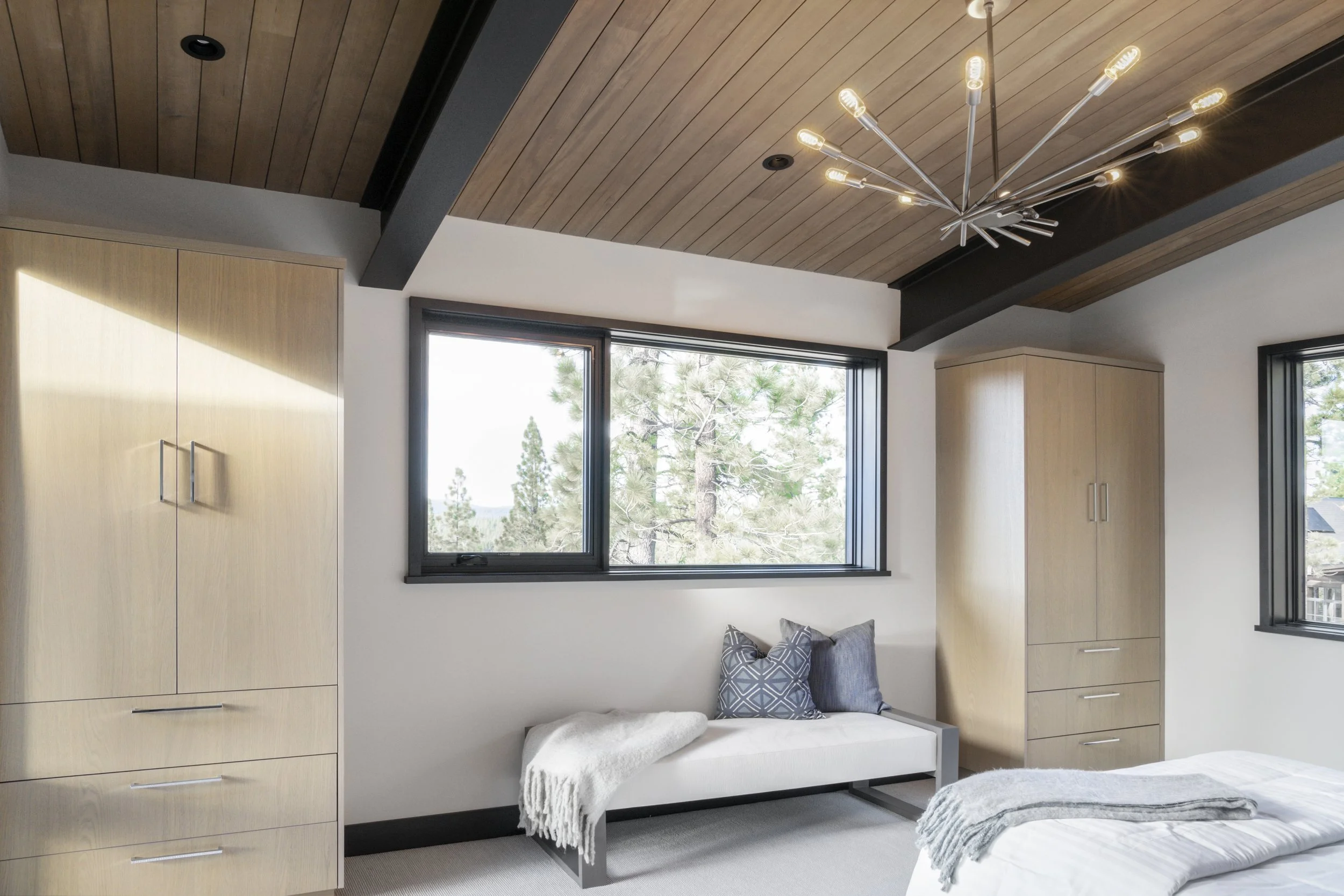 A bedroom with a wooden ceiling featuring a modern chandelier, white walls, black window frames, a white bed with a gray throw, and a small white bench with pillows and a throw blanket.