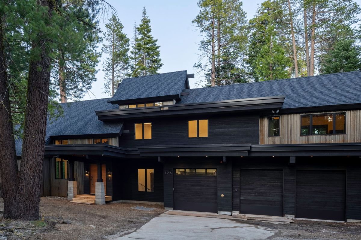 A modern black two-story house with a sloped roof, located in a wooded area with tall trees. The house has multiple windows, a small front porch, and a garage with three doors.