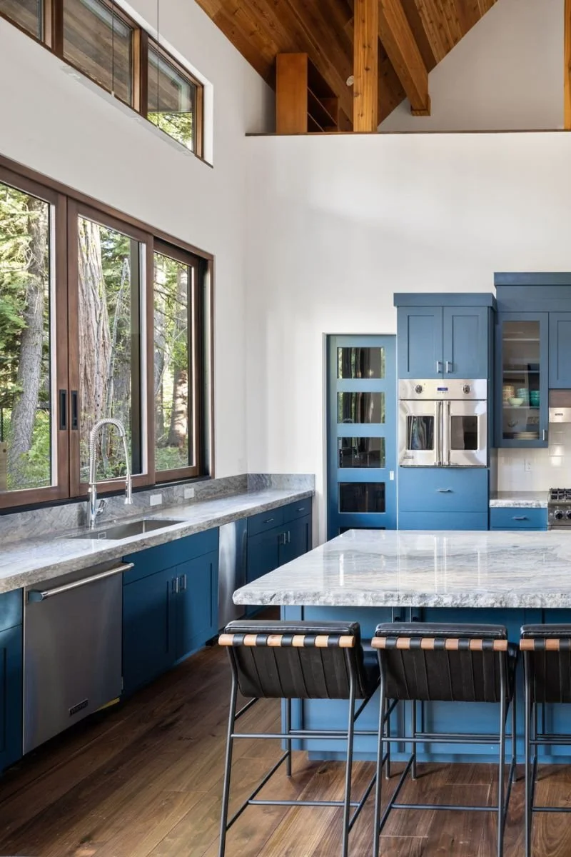 Modern kitchen with blue cabinets, white marble countertops, large windows showing outdoor trees, and wooden ceiling beams.