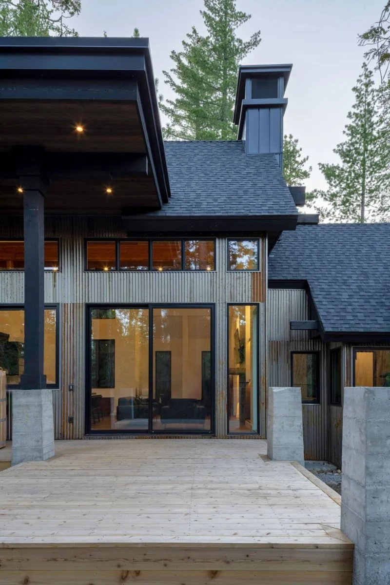 Modern house with large glass sliding doors, wooden siding, and a wooden deck, surrounded by tall trees.