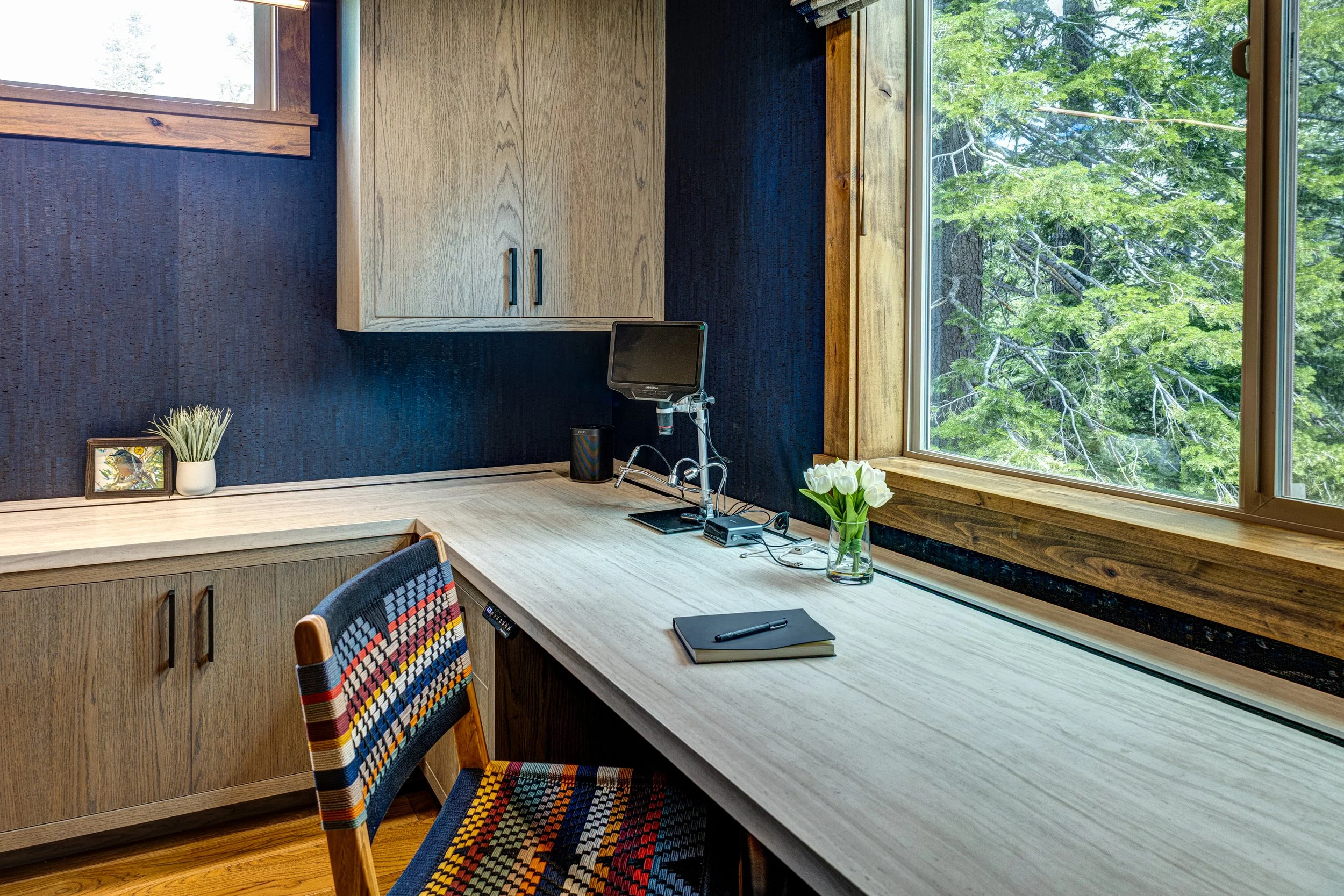 Home office with wooden desk and cabinet, colorful woven chair, small plant, monitor, notebook, and flowers, overlooking lush green trees through large window.