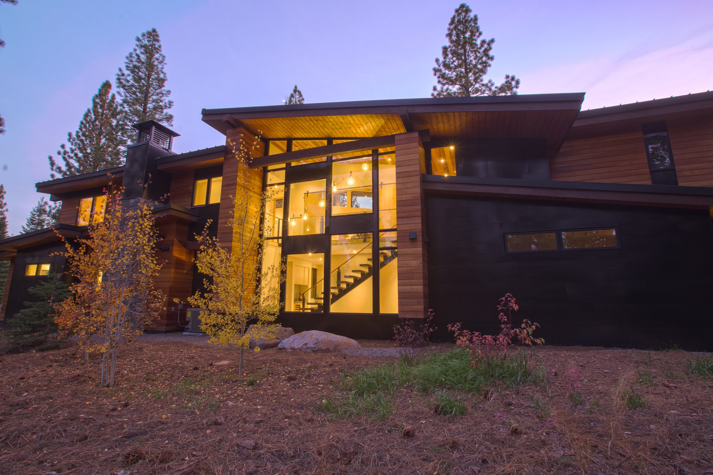 Modern house with large glass windows illuminated with warm interior lighting, surrounded by trees and natural landscape, during dusk.