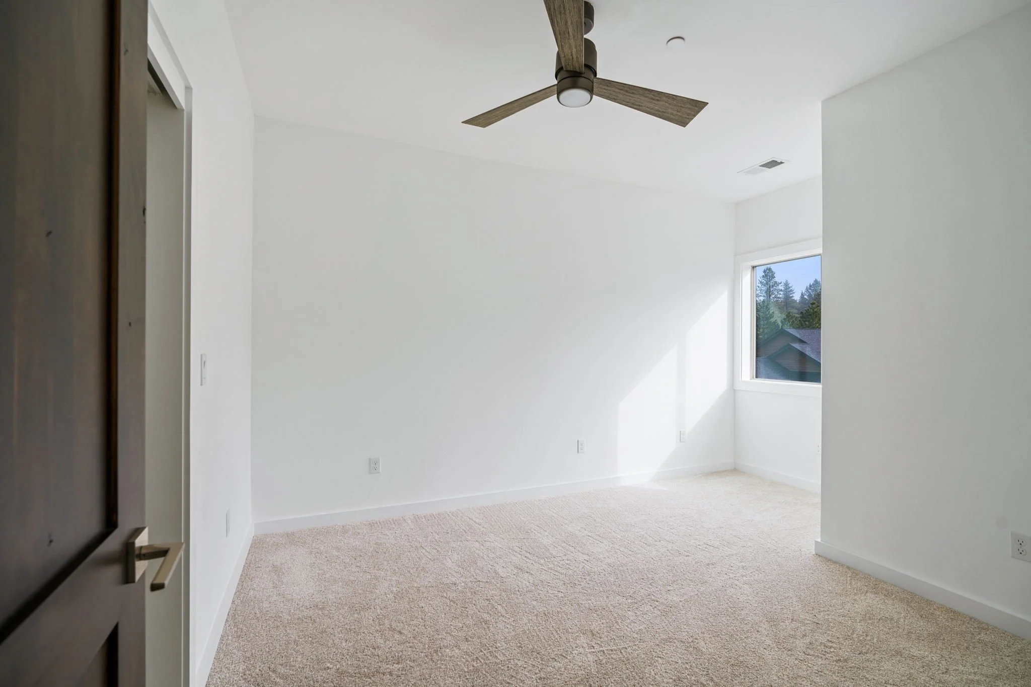 Empty room with beige carpet, white walls, a window showing trees outside, and a ceiling fan with wooden blades.