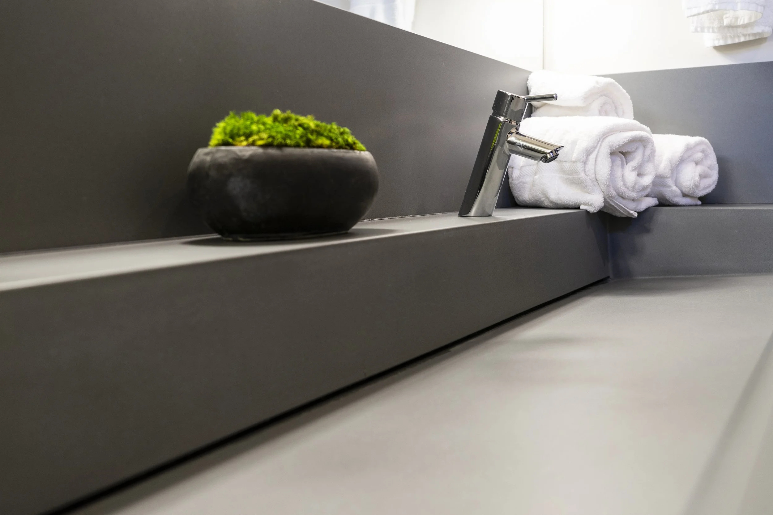 Bathroom countertop with a black flower pot, white rolled towels, modern chrome faucet, gray back splash, and white wall.
