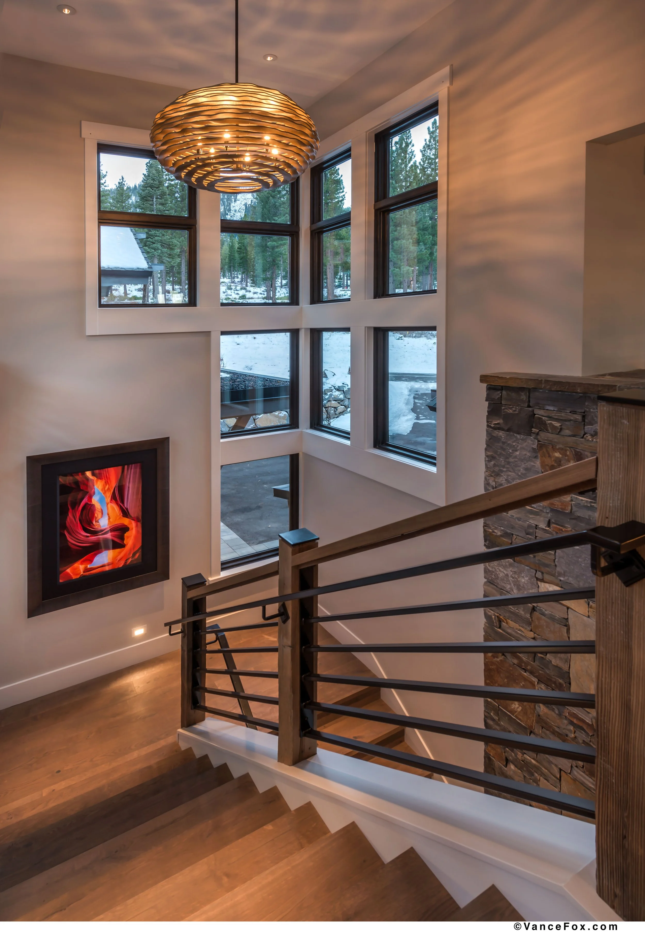 Interior view of a home staircase with wooden steps and black metal railing, overlooking a double-height living area with large windows showing a snowy outdoor landscape, a modern fireplace, and a wooden and stone decor accent.