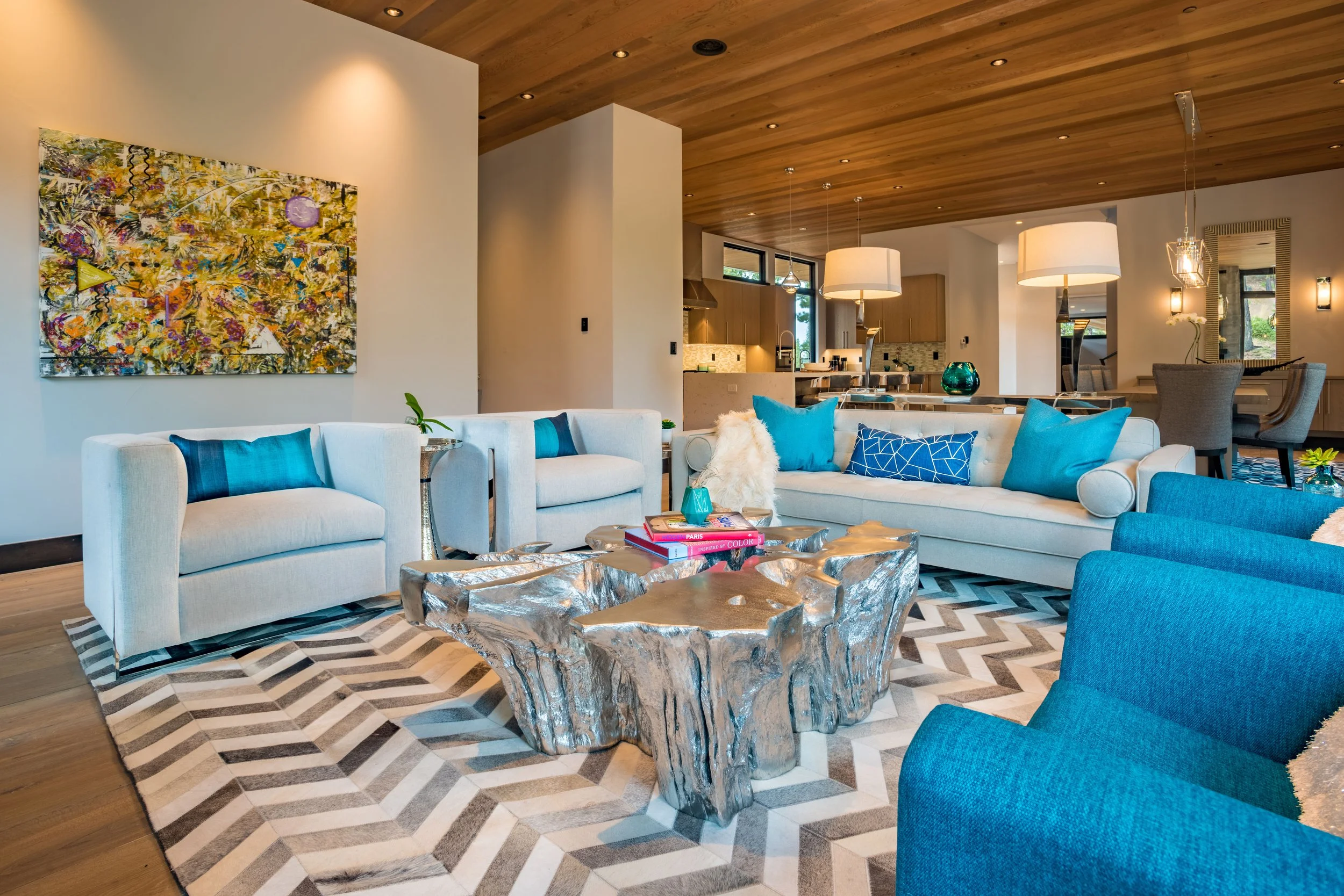 Modern living room with white and blue sofas, a metallic sculptural coffee table, patterned rug, colorful artwork on the wall, and a cozy open kitchen in the background with pendant lighting.