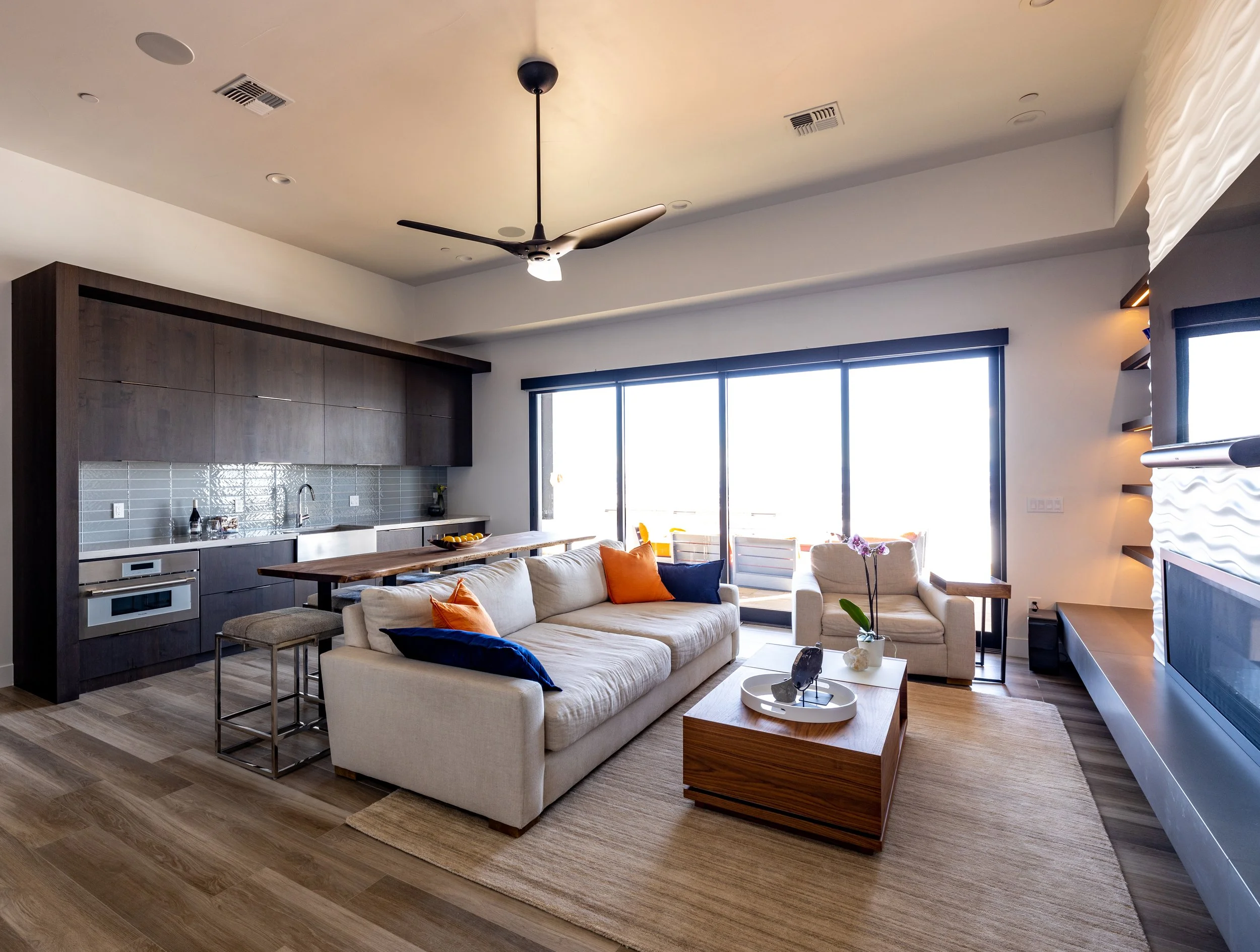 Living room with a beige sofa, armchair, wooden coffee table, and decorative pillows, adjacent to a modern kitchen with dark cabinets, gray backsplash, and large sliding glass doors leading to an outdoor balcony.