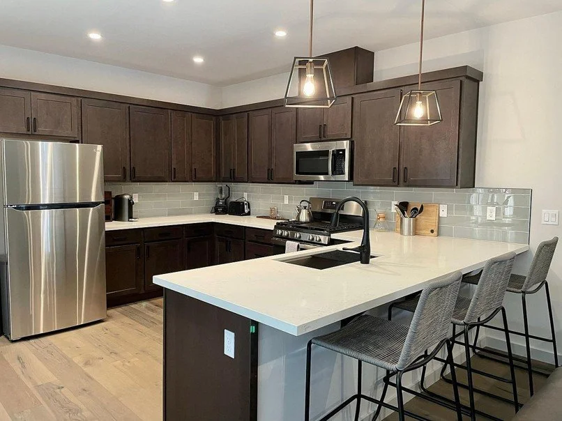 Modern kitchen with dark wood cabinets, stainless steel refrigerator, microwave, and oven, gray subway tile backsplash, white countertops, and gray bar stools at an island with a black sink and faucet.