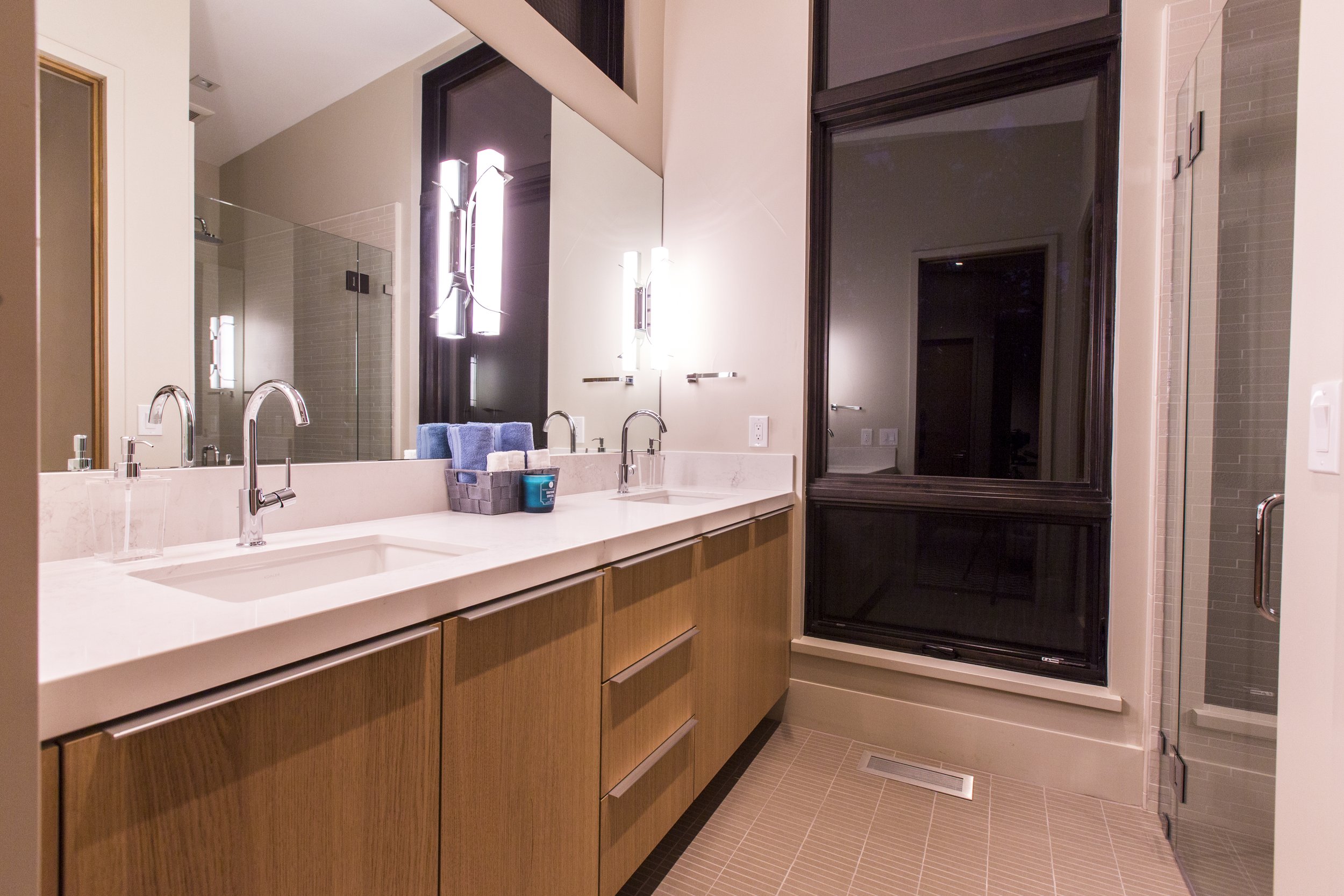 Modern bathroom with dual sinks, large mirror, wooden vanity, black-framed window, walk-in shower, and neutral tones.