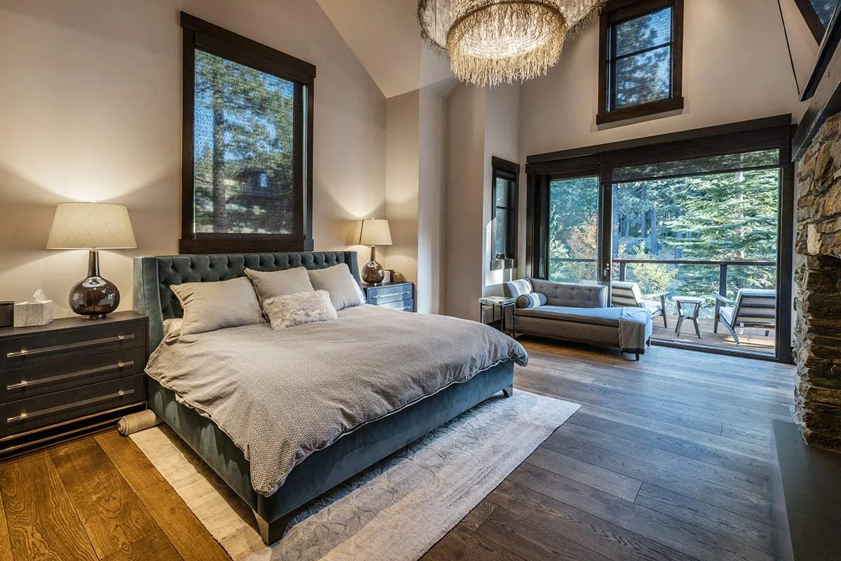 A spacious bedroom with a large bed featuring a tufted headboard, two nightstands with lamps, a bench near the sliding glass door, and a balcony overlooking trees.