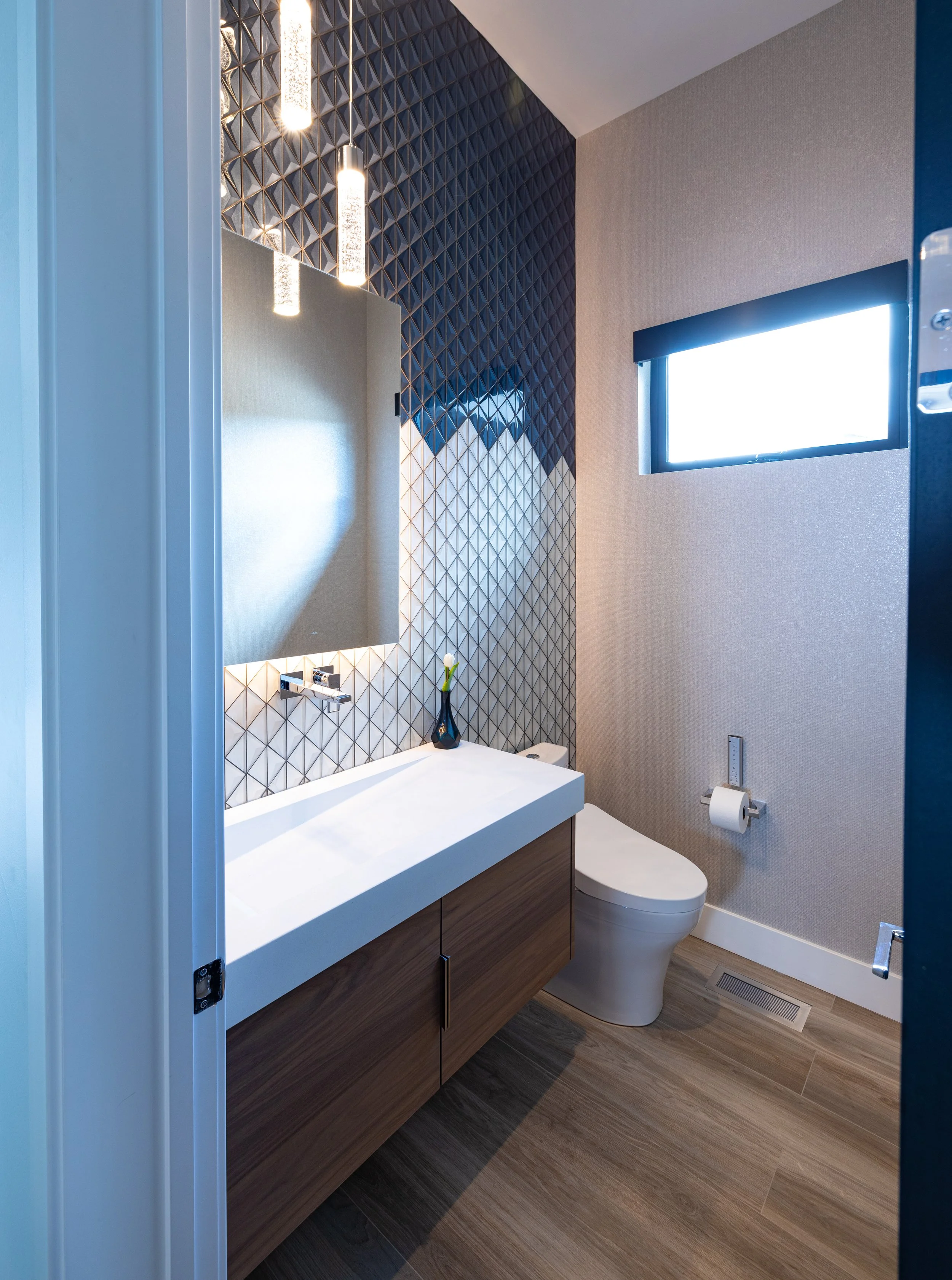 Modern bathroom with a wooden vanity, a large beige mirror, three hanging lights, a geometric black and white tile accent wall, a small window, and a white toilet.