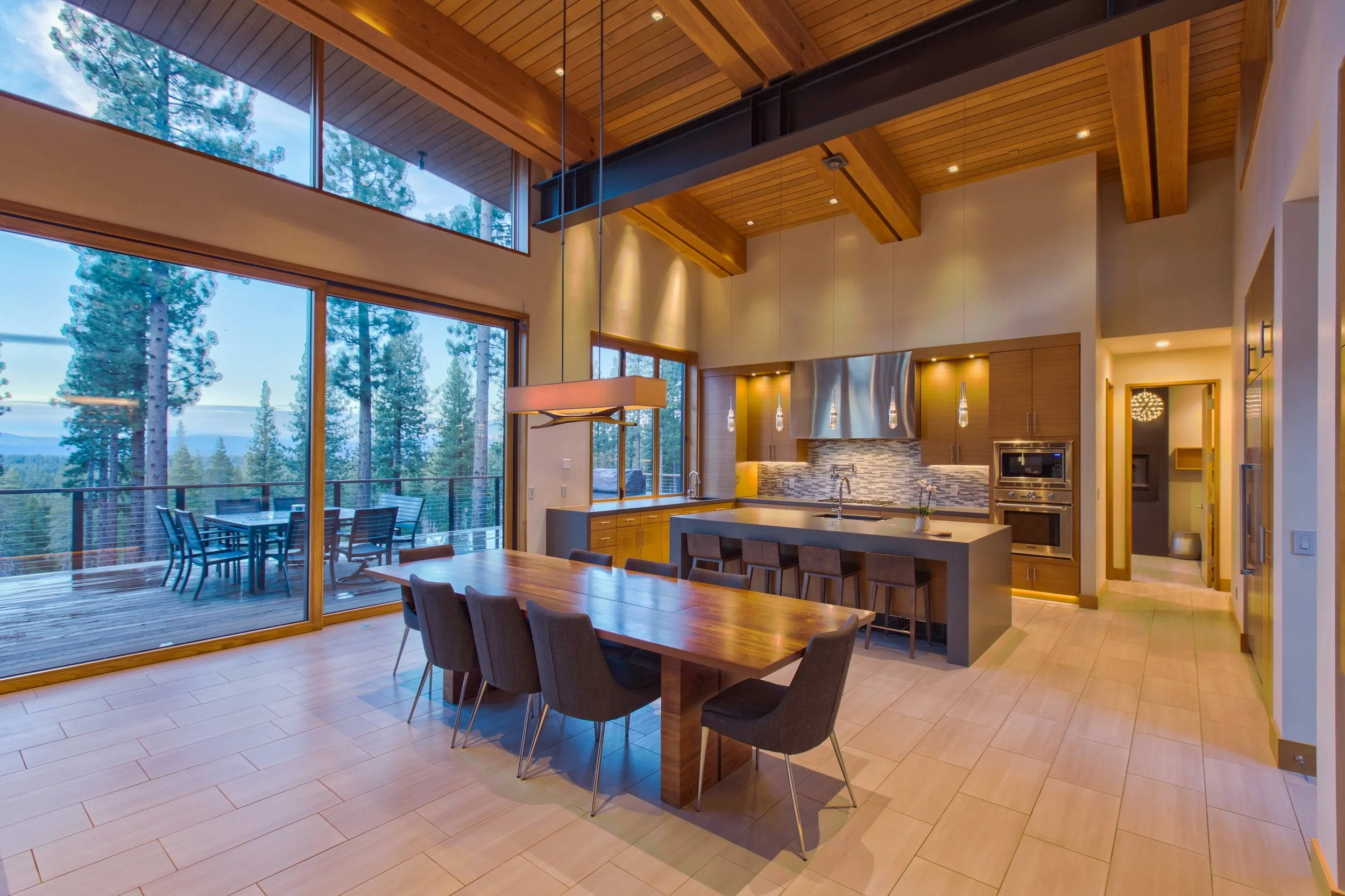Interior view of a modern open-concept kitchen and dining area with large windows showing a scenic forested landscape, wooden ceiling beams, and a balcony with an outdoor dining set.