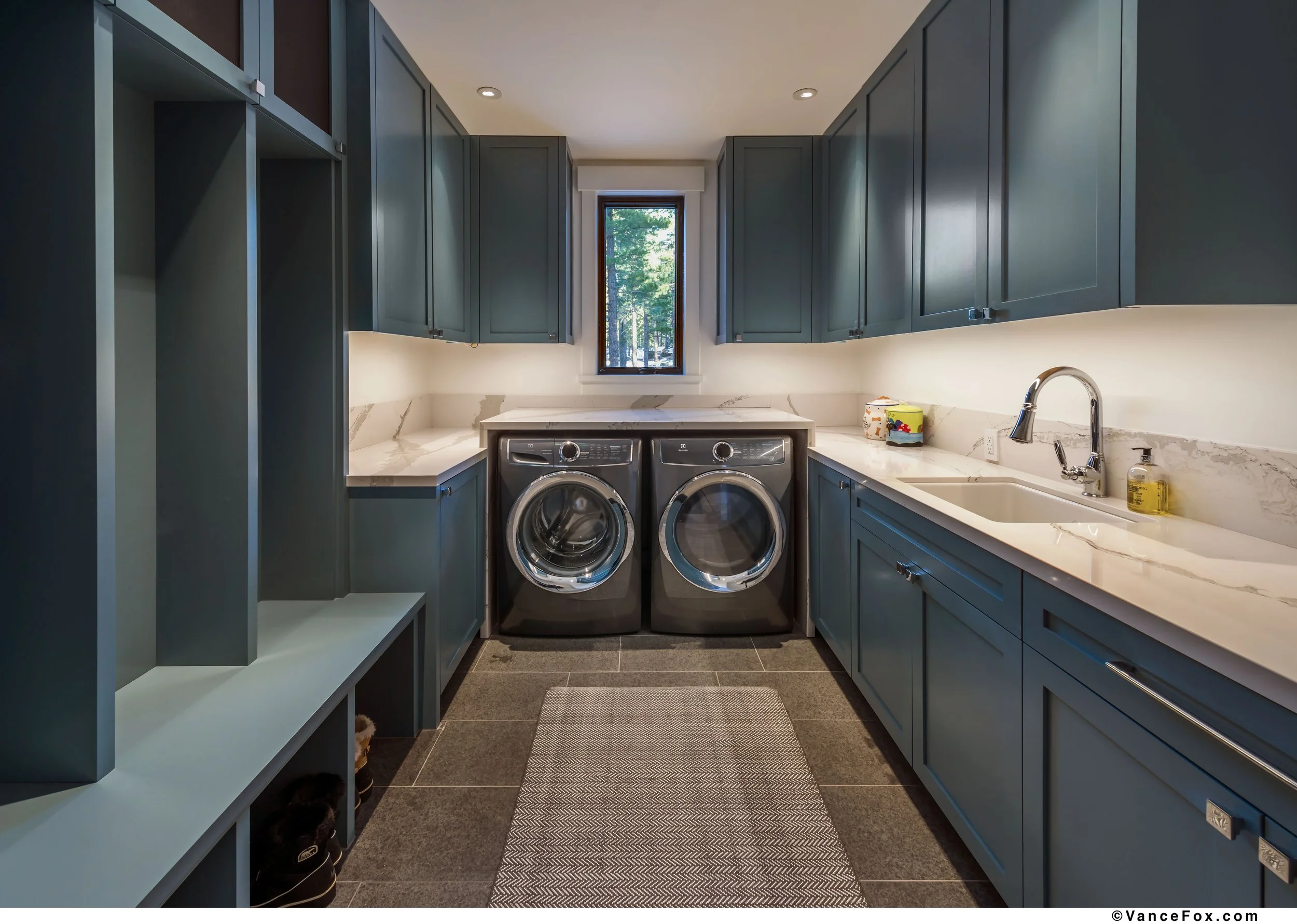 Laundry room with blue cabinets, quartz countertops, front-loading washing machine and dryer, sink, and small window showing trees outside.