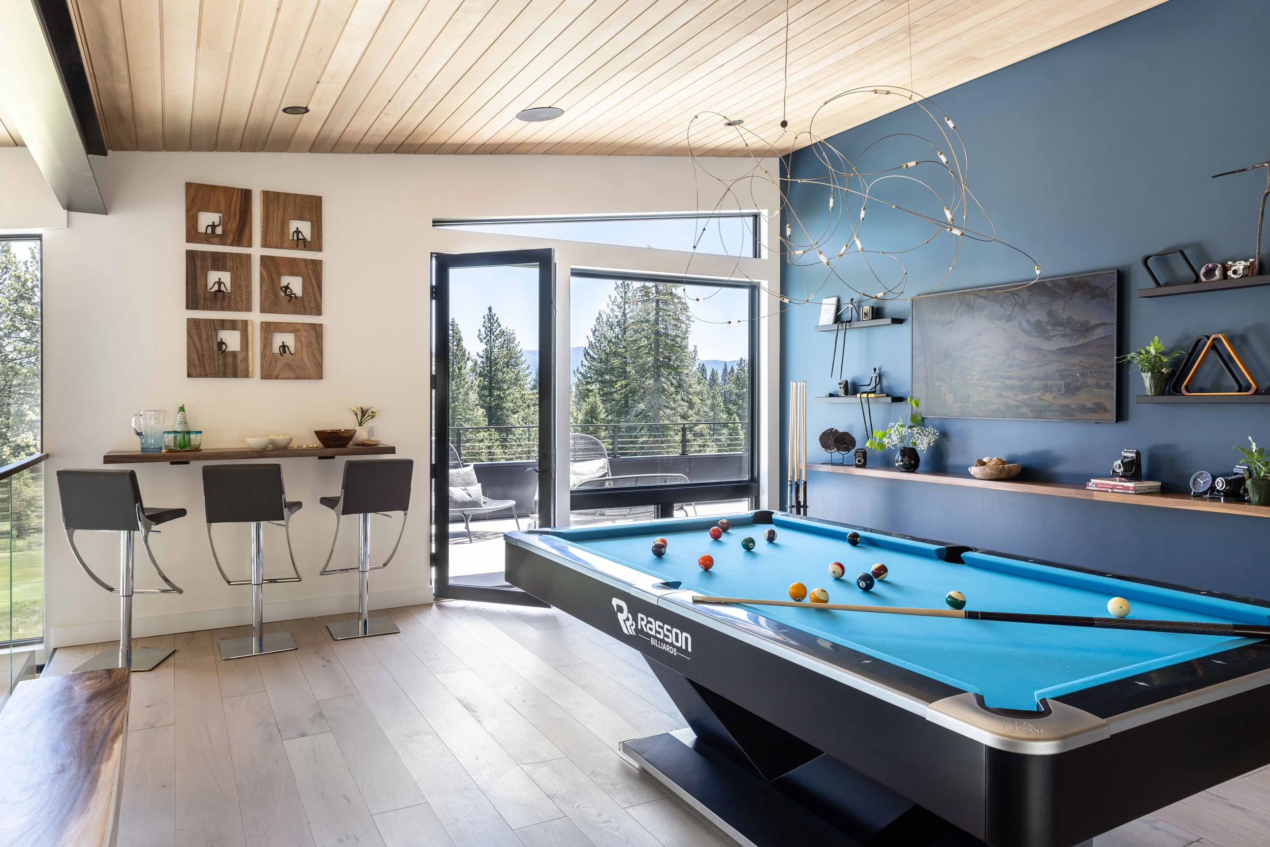 A modern game room with a billiards table, a wall-mounted TV, shelves with decor, a small bar area with three stools, and a balcony with outdoor seating overlooking a forested landscape.