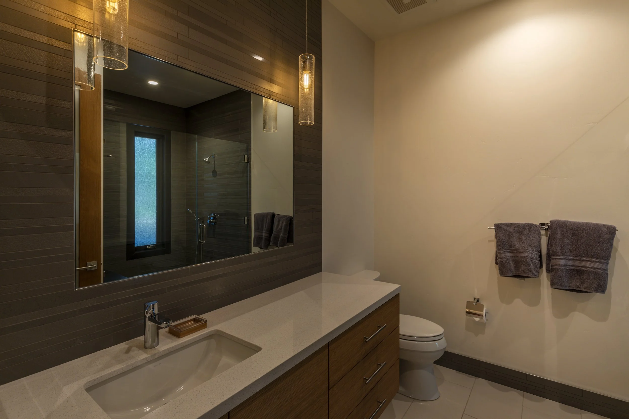 Modern bathroom with a beige countertop, wooden cabinets, a large mirror, and a toilet. Towels hang on a rail, and there is a shower visible through a window in the reflection.